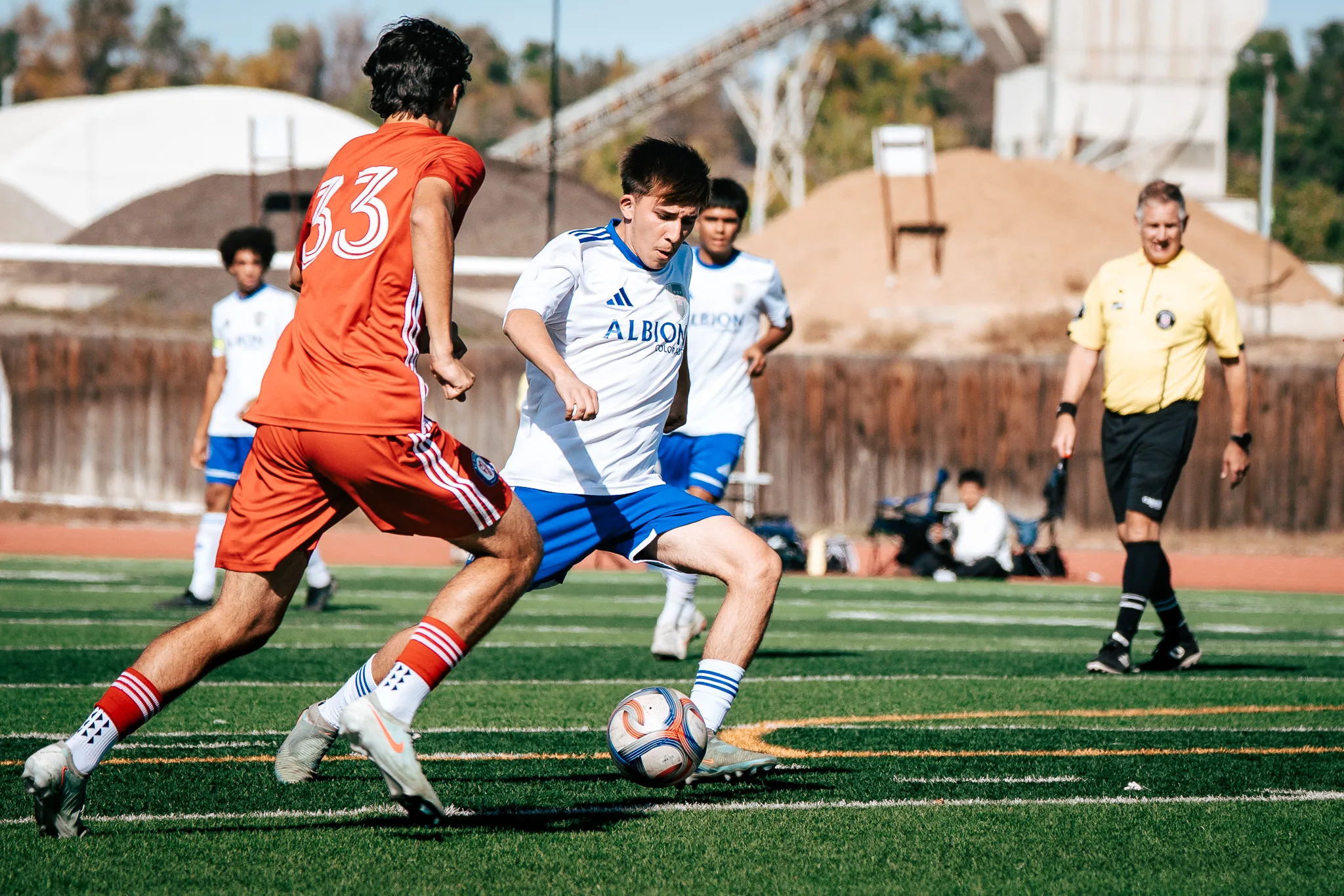 An Albion SC Colorado player dribbles at AC River number 33 as two teammates follow and the referee in yellow watches with a smile