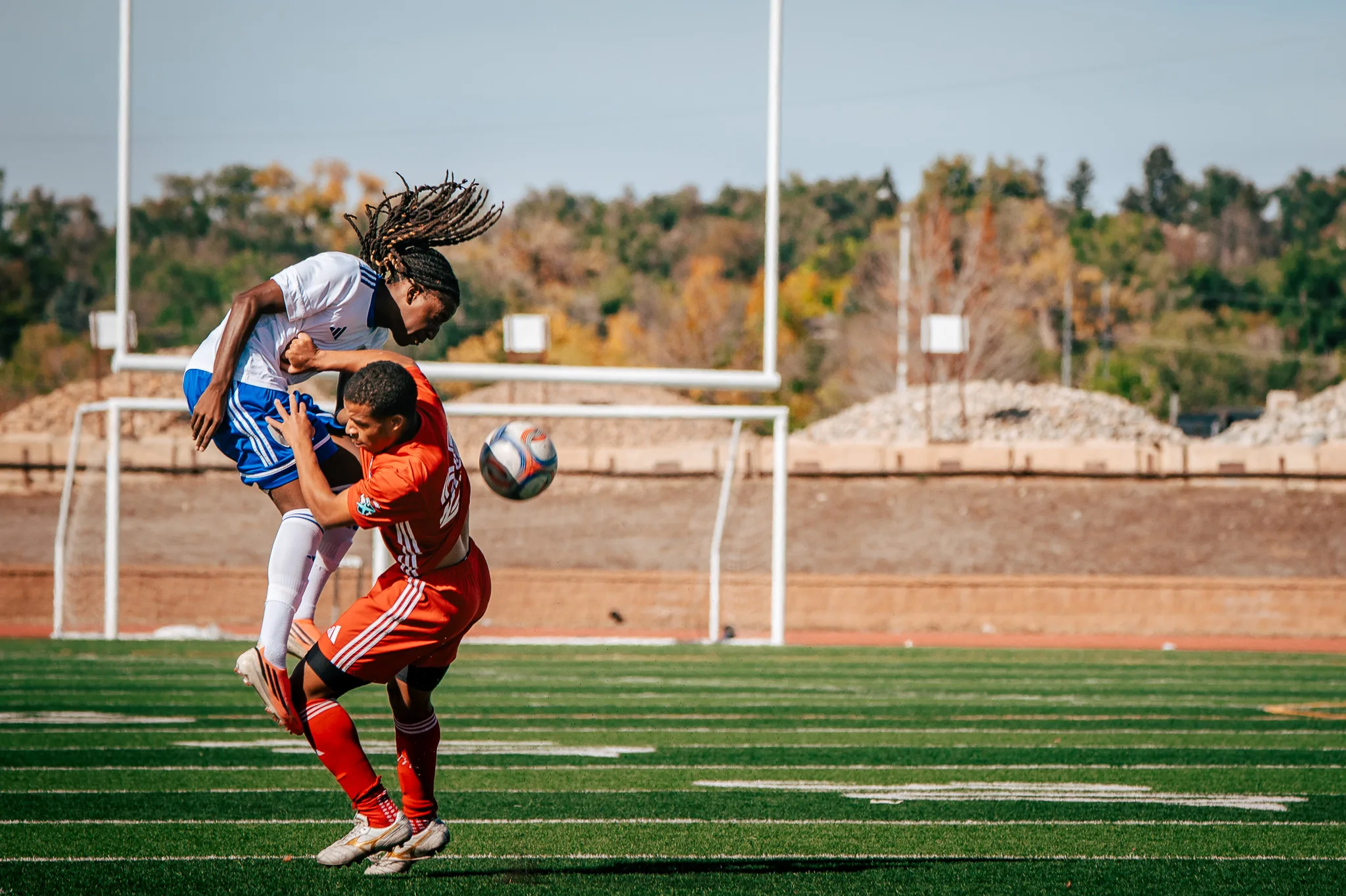 In bright sun, an Albion SC Colorado player with long locs goes up over an AC River player as the ball flies past them, goalposts and autumn trees behind