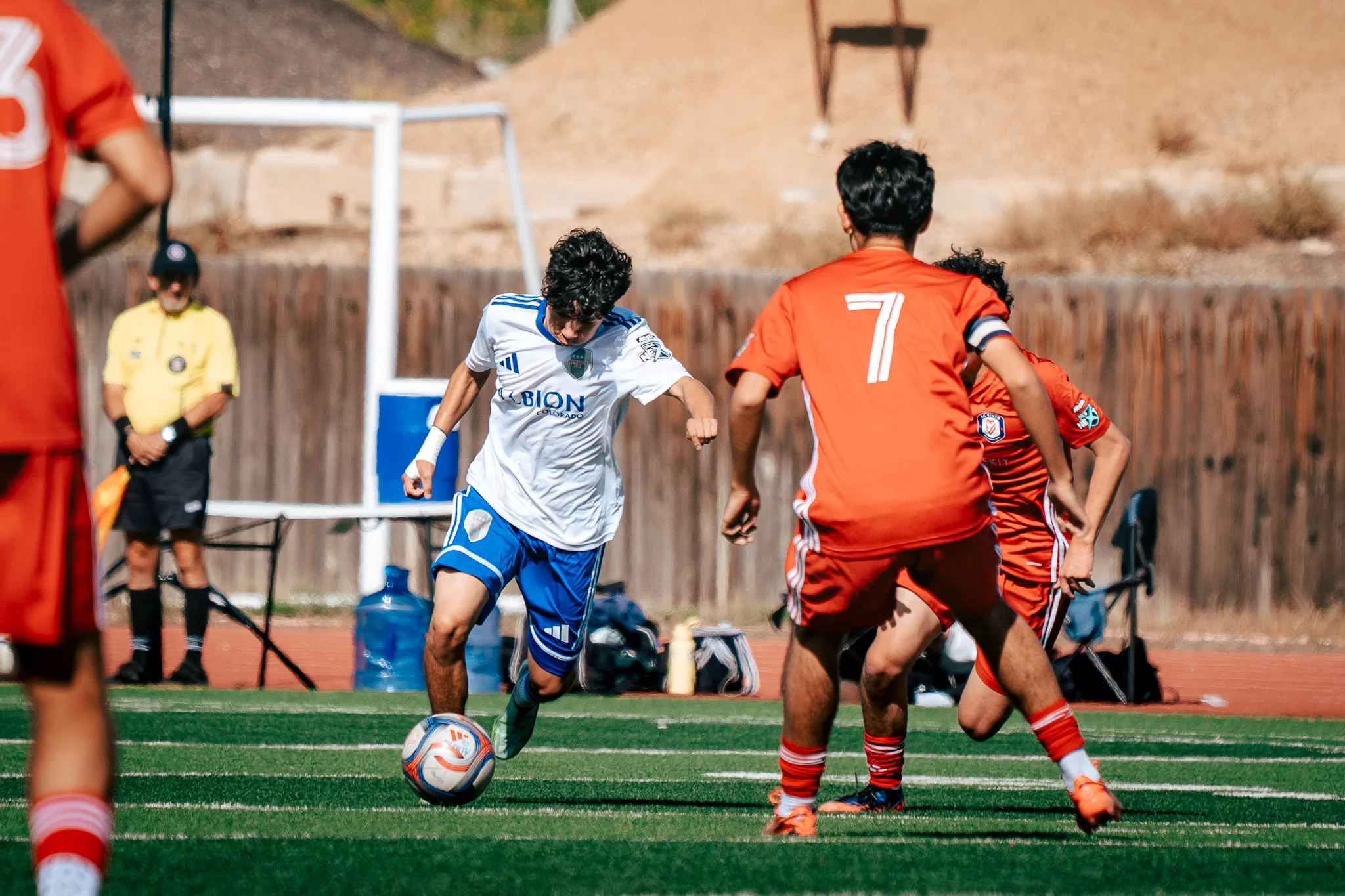 An Albion SC Colorado player dribbles toward AC River number 7, in a captain's armband, as an assistant referee in yellow watches from the sideline