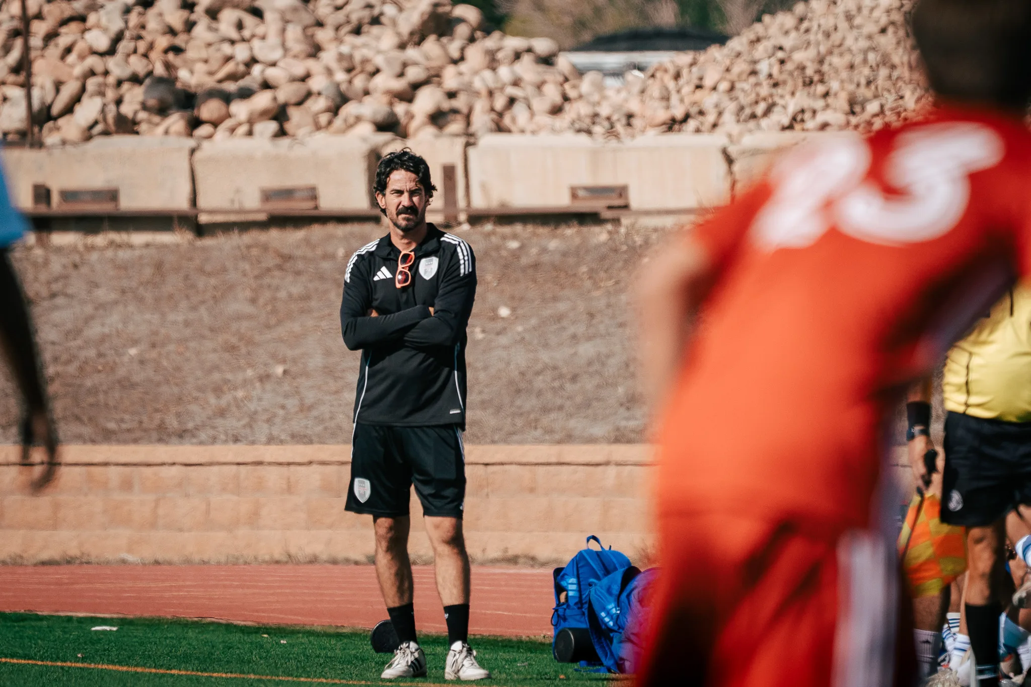 An Albion SC Colorado coach stands on the sideline with his arms crossed and sunglasses hooked on his shirt as a blurred AC River player passes in the foreground
