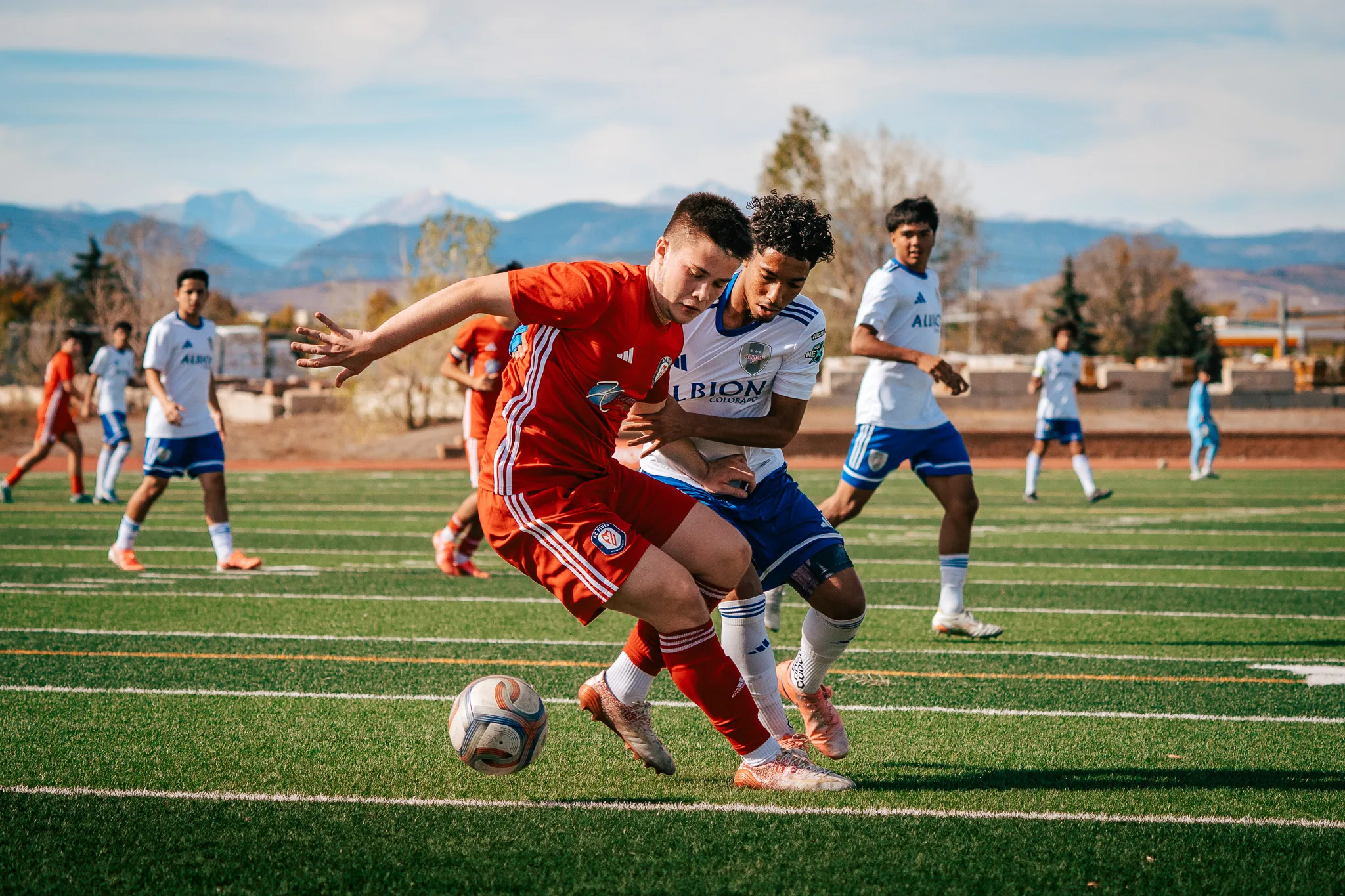 An AC River player holds off an Albion SC Colorado player at the ball, snowy mountains behind