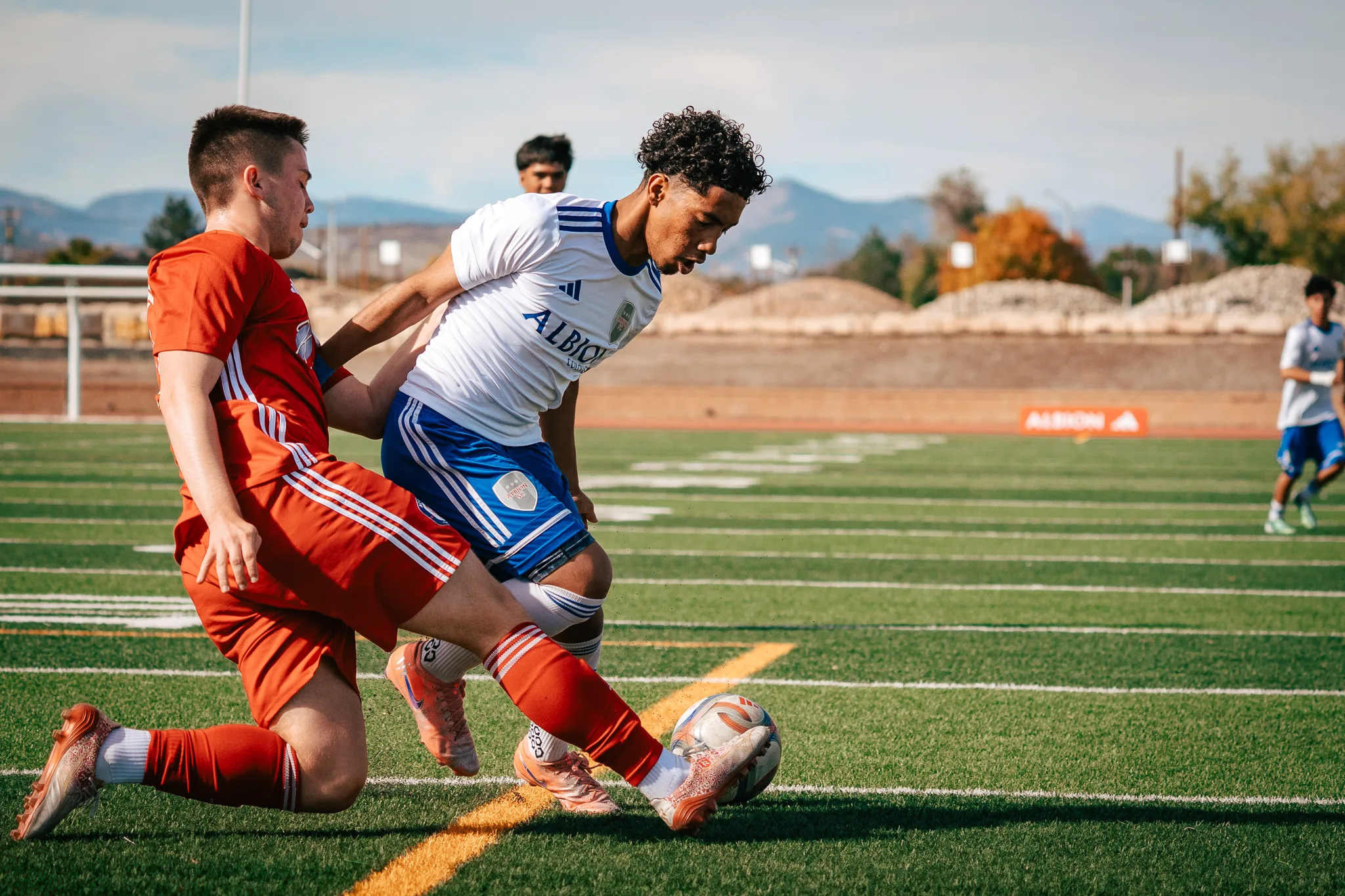 An AC River player slides in at the ball as an Albion SC Colorado player steps over it, mountains behind