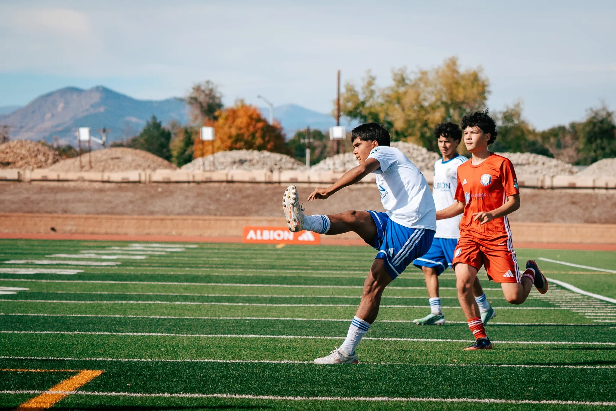 An Albion SC Colorado player swings his leg high after a kick as a teammate and an AC River player run behind, a mountain and autumn trees in the distance