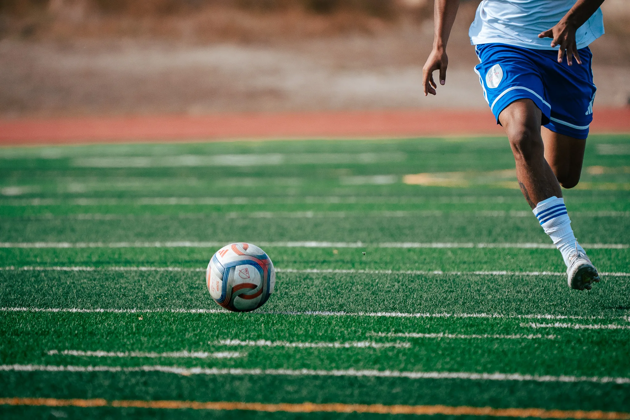 Low to the turf, an Albion SC Colorado player in blue shorts runs onto a rolling ball