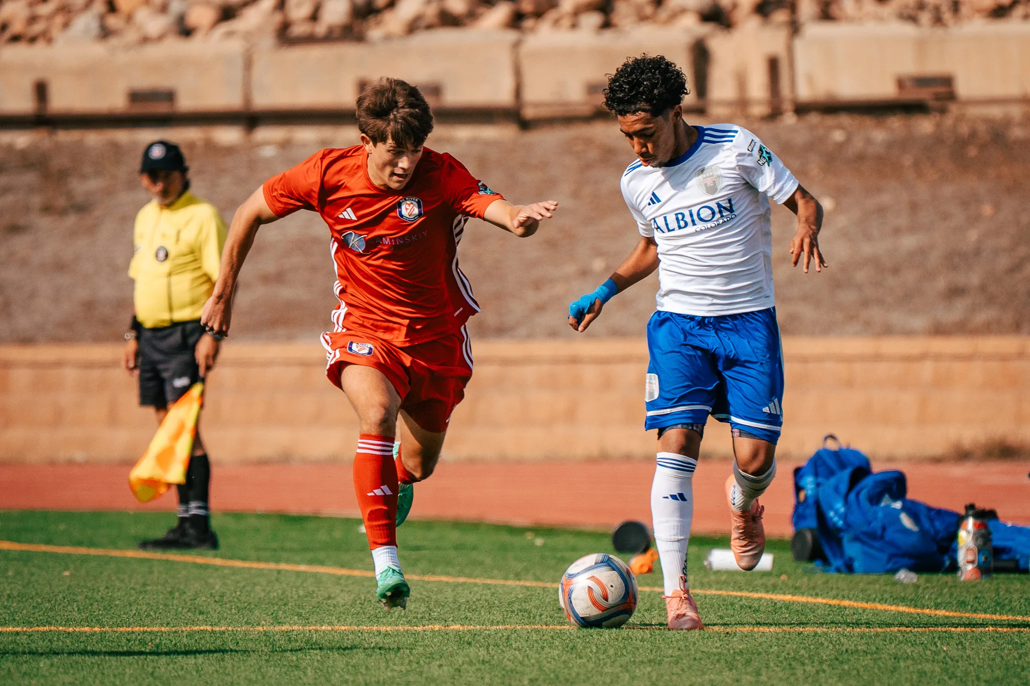 An AC River player and an Albion SC Colorado player with blue wrist tape race for the ball as an assistant referee holds his flag on the sideline
