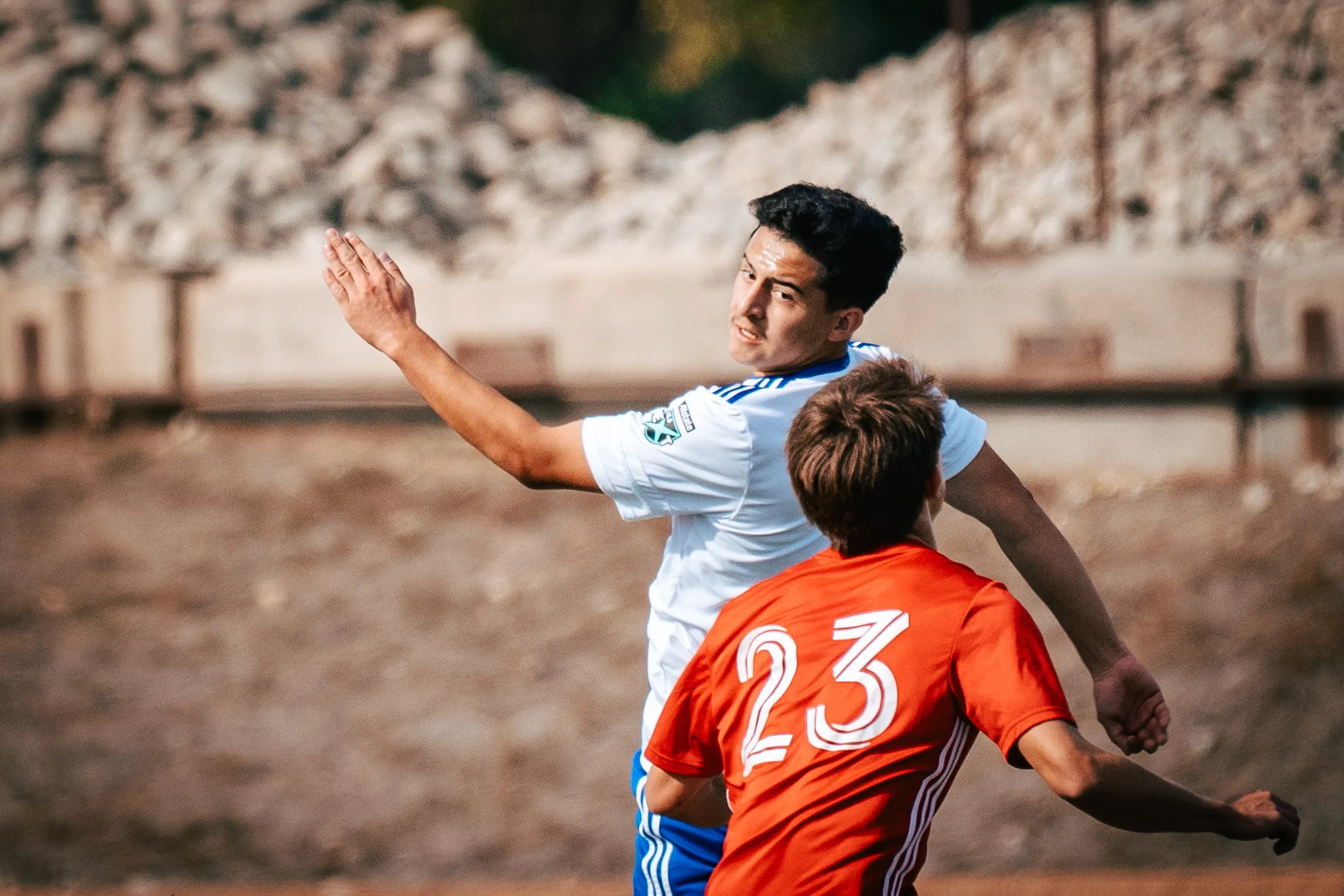 An Albion SC Colorado player throws an arm up as AC River number 23 leans into him, a hill of rocks behind