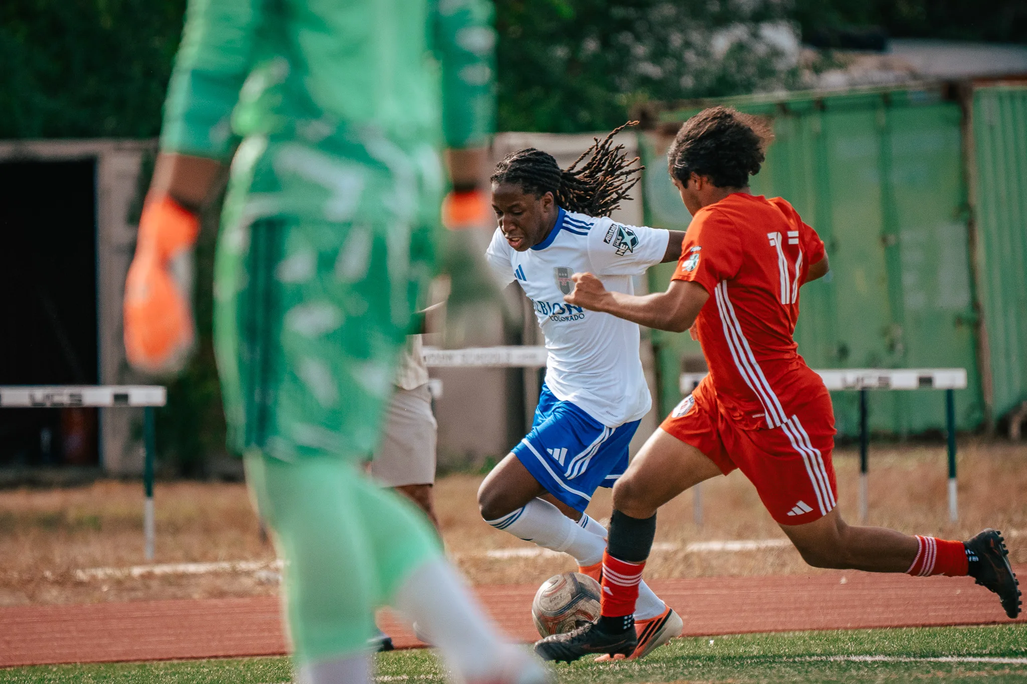 An Albion SC Colorado player with long locs drives at the ball past an AC River player, the AC River goalkeeper in green blurred in the foreground