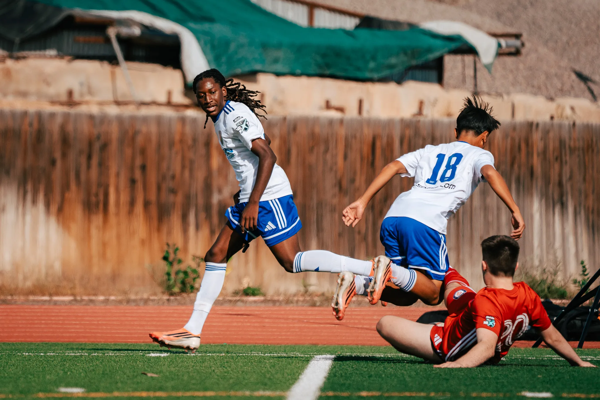 Albion SC Colorado number 18 hurdles AC River number 20, down on the turf, as an Albion teammate with long locs looks back