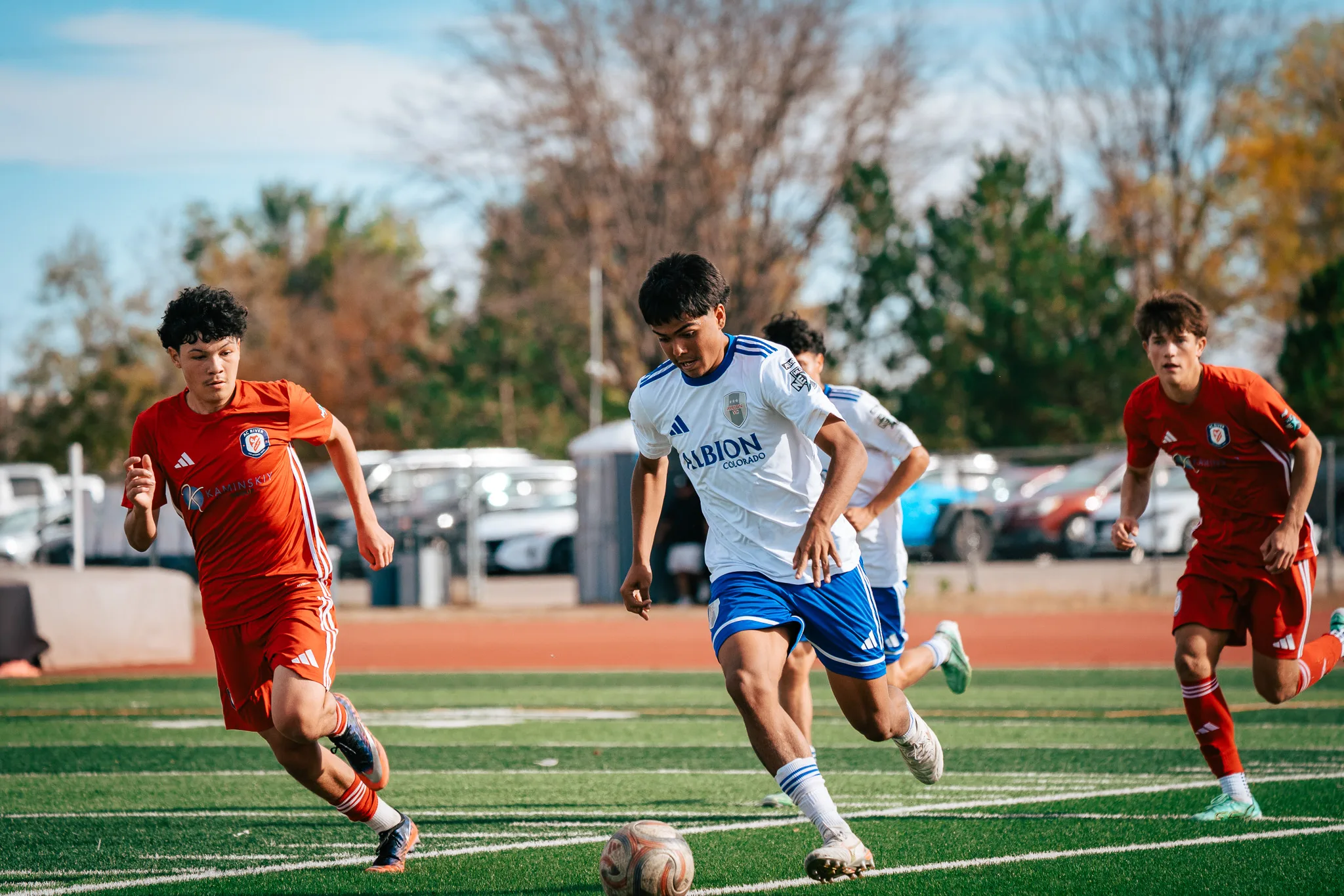 An Albion SC Colorado player carries the ball between two AC River players, a teammate close behind and parked cars beyond the track
