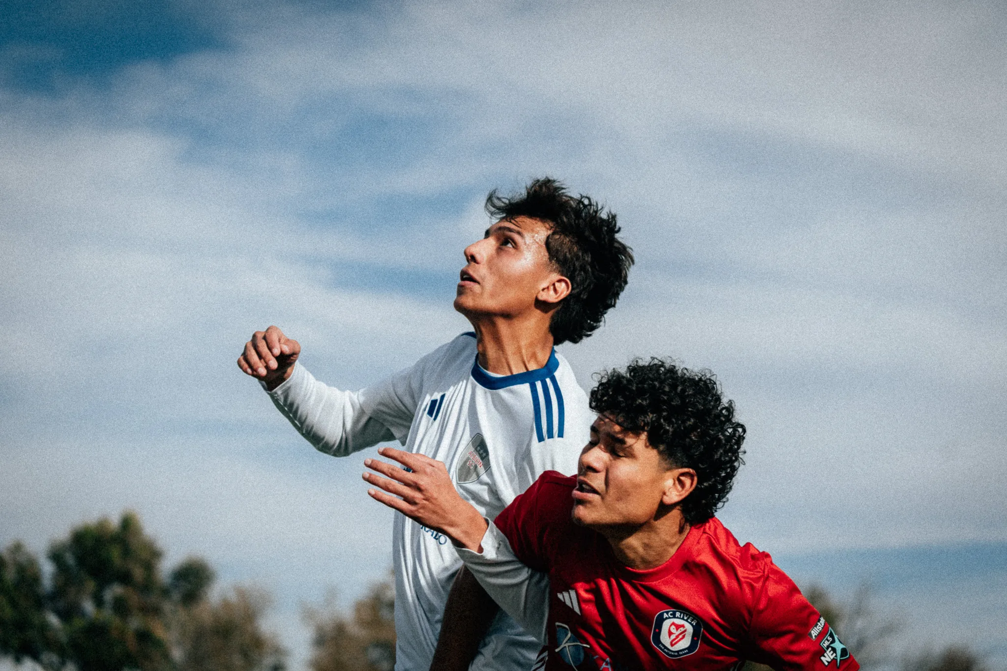 Against a blue sky, an Albion SC Colorado player and an AC River player look up together for a high ball