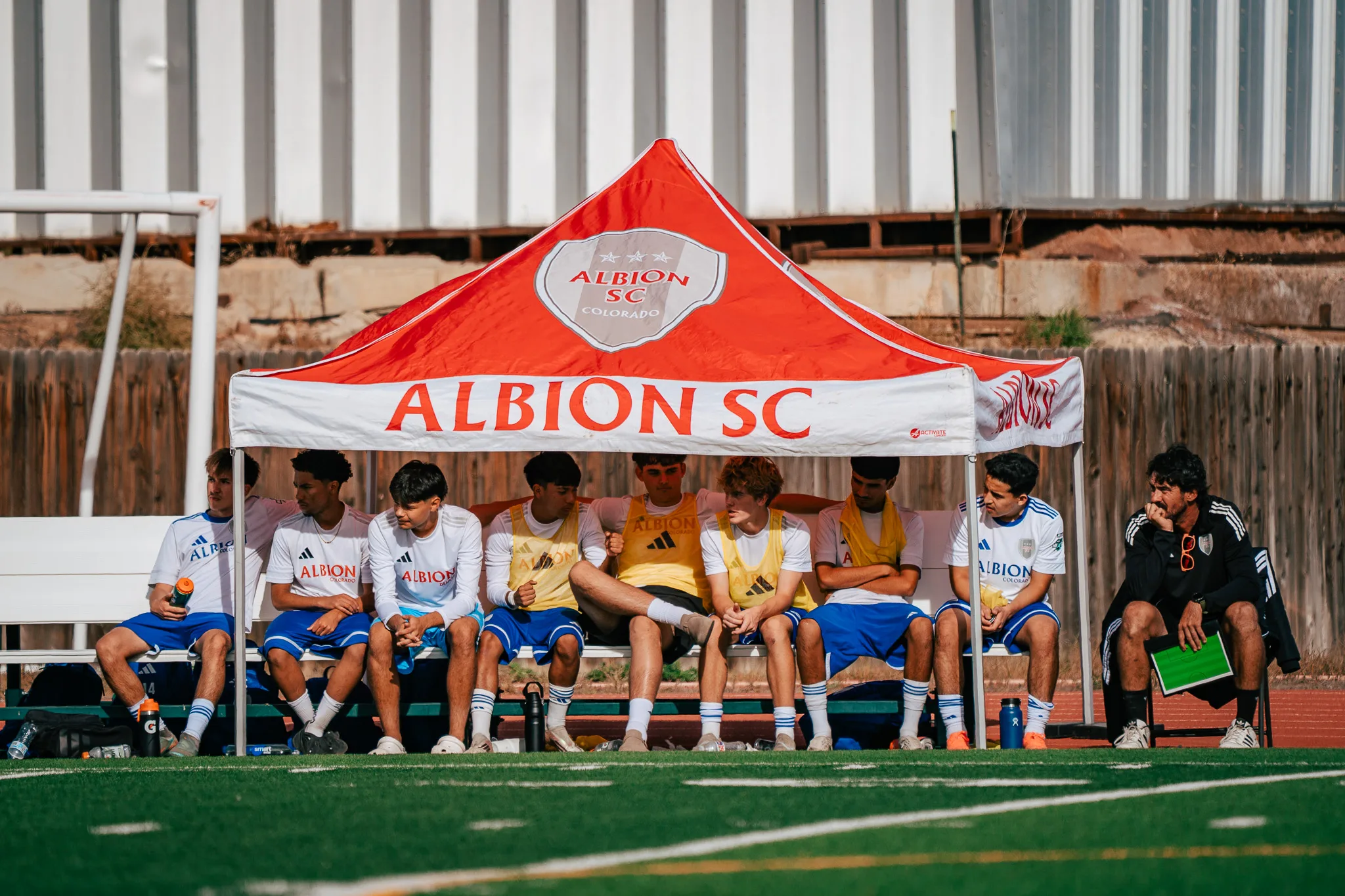 Albion SC Colorado substitutes sit on the bench under a red Albion SC tent, some in yellow pinnies, a coach with a green clipboard at the end