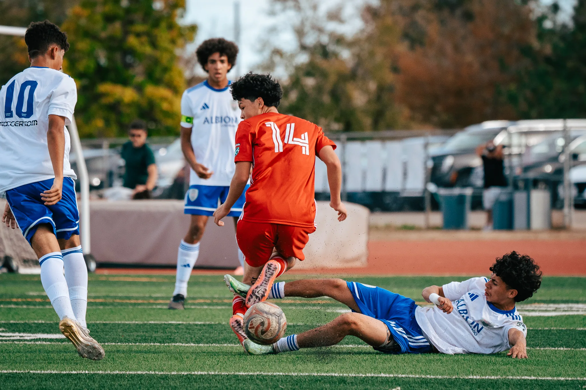 AC River number 14 rides a sliding tackle from an Albion SC Colorado player as Albion number 10 and a teammate in a captain's armband look on