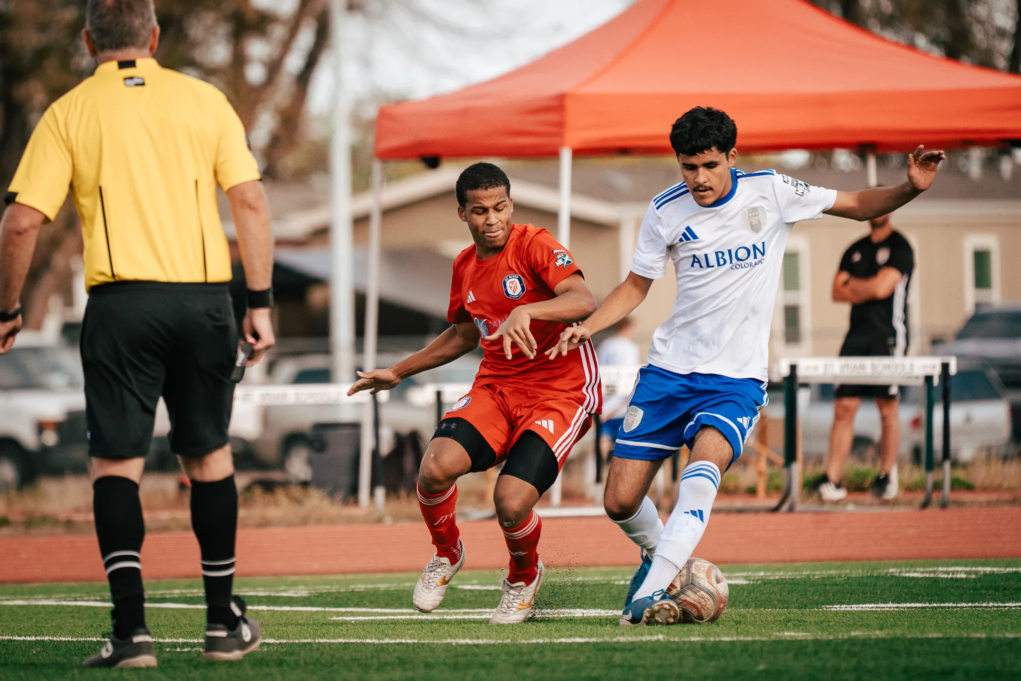 An Albion SC Colorado player steps past an AC River player with the ball as the referee watches from behind, a red canopy across the track