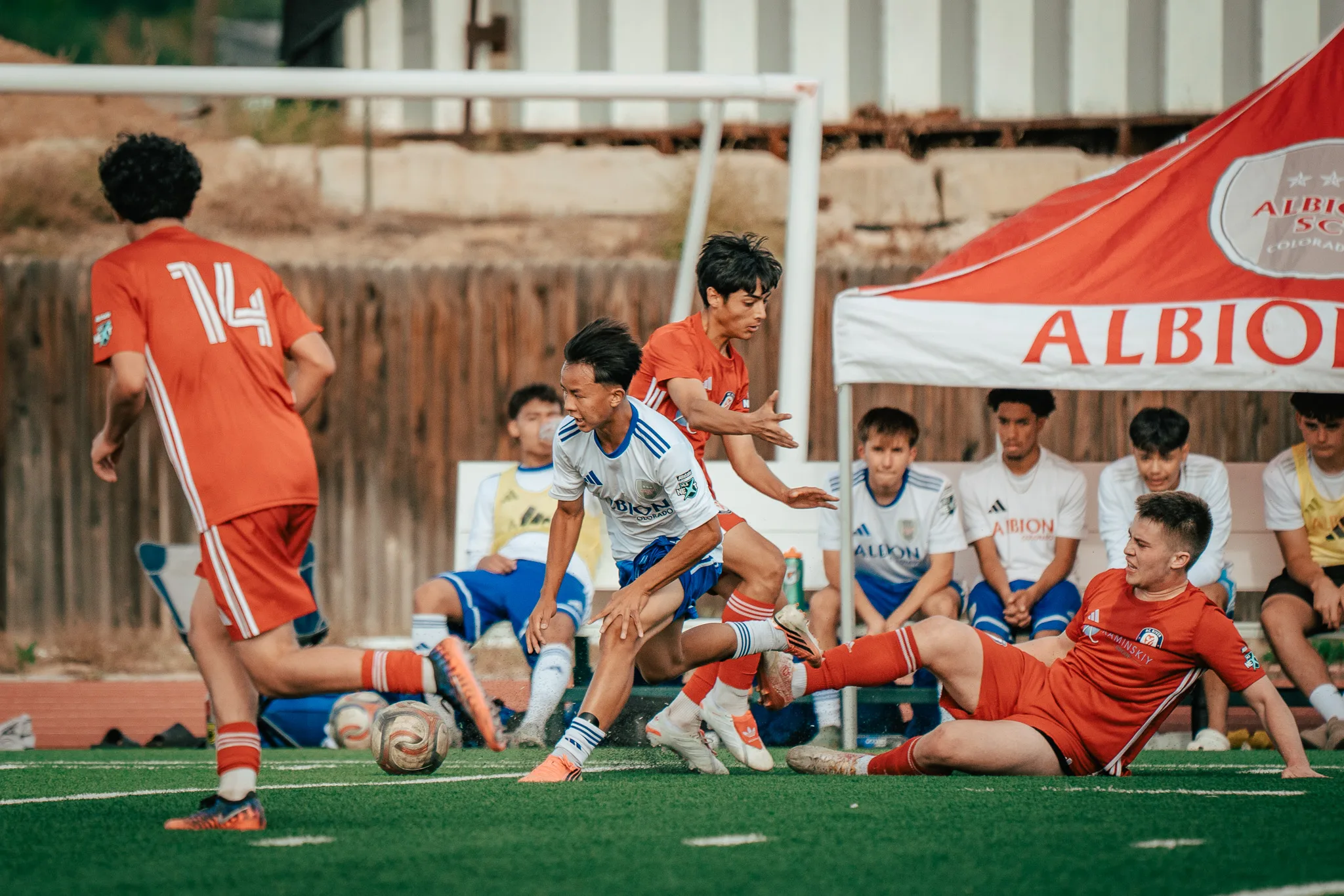 An Albion SC Colorado player cuts between AC River players, number 14 among them and one down on the turf, in front of the Albion bench and its red tent