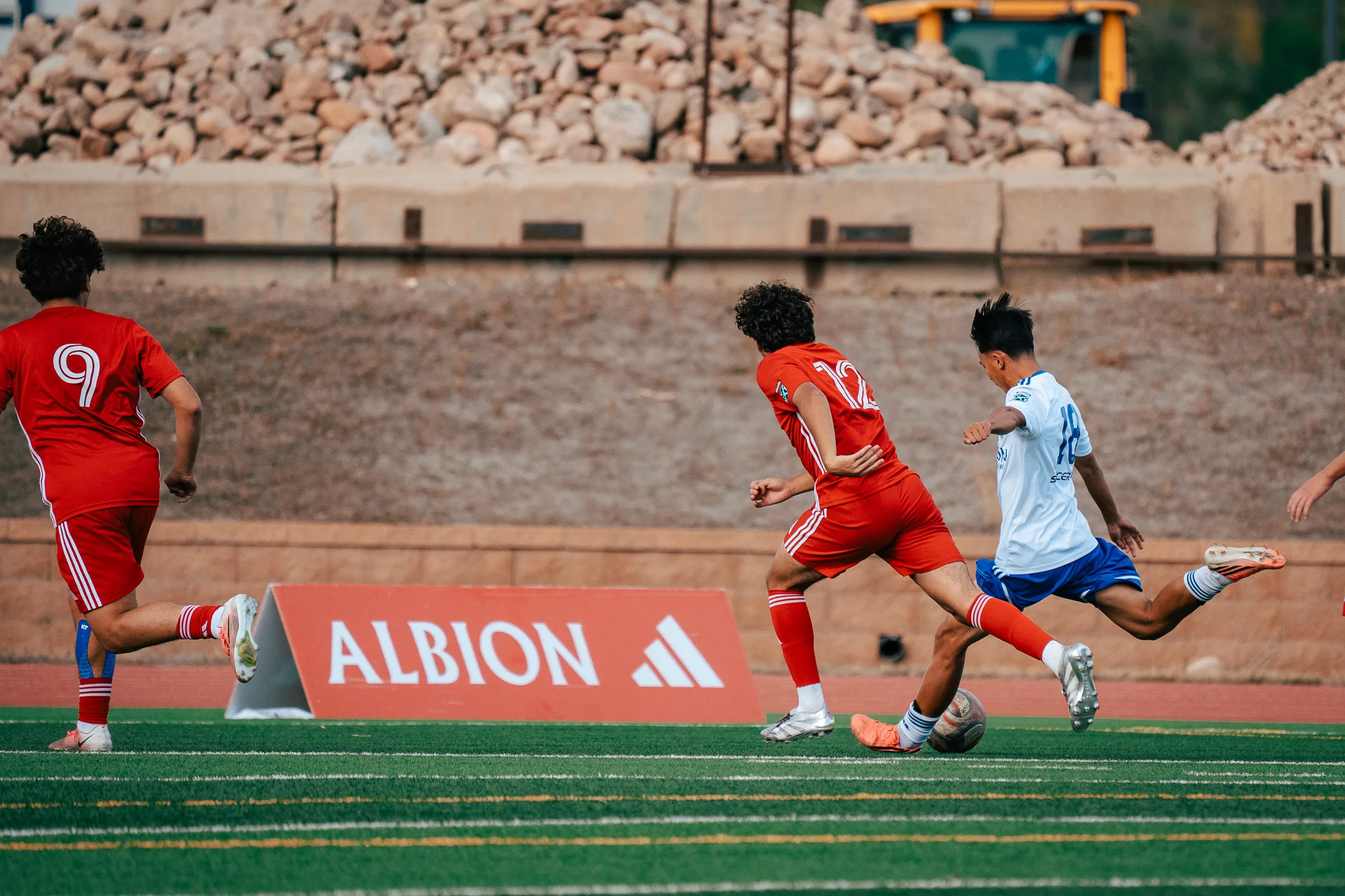Albion SC Colorado number 18 strikes the ball as AC River number 12 stretches to block and AC River number 9 runs in, a red Albion sign behind