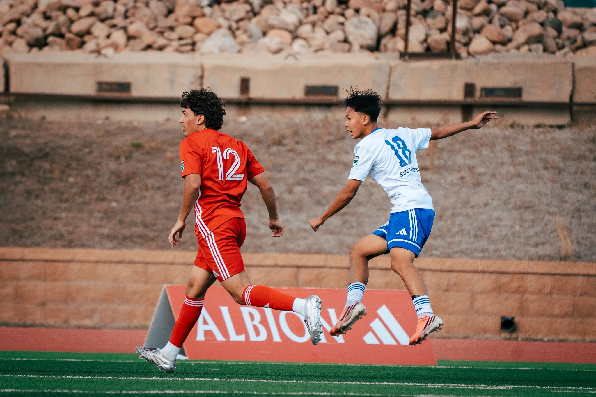 Albion SC Colorado number 18 leaps with one arm out beside AC River number 12, both turned to follow the ball, a red Albion sign behind
