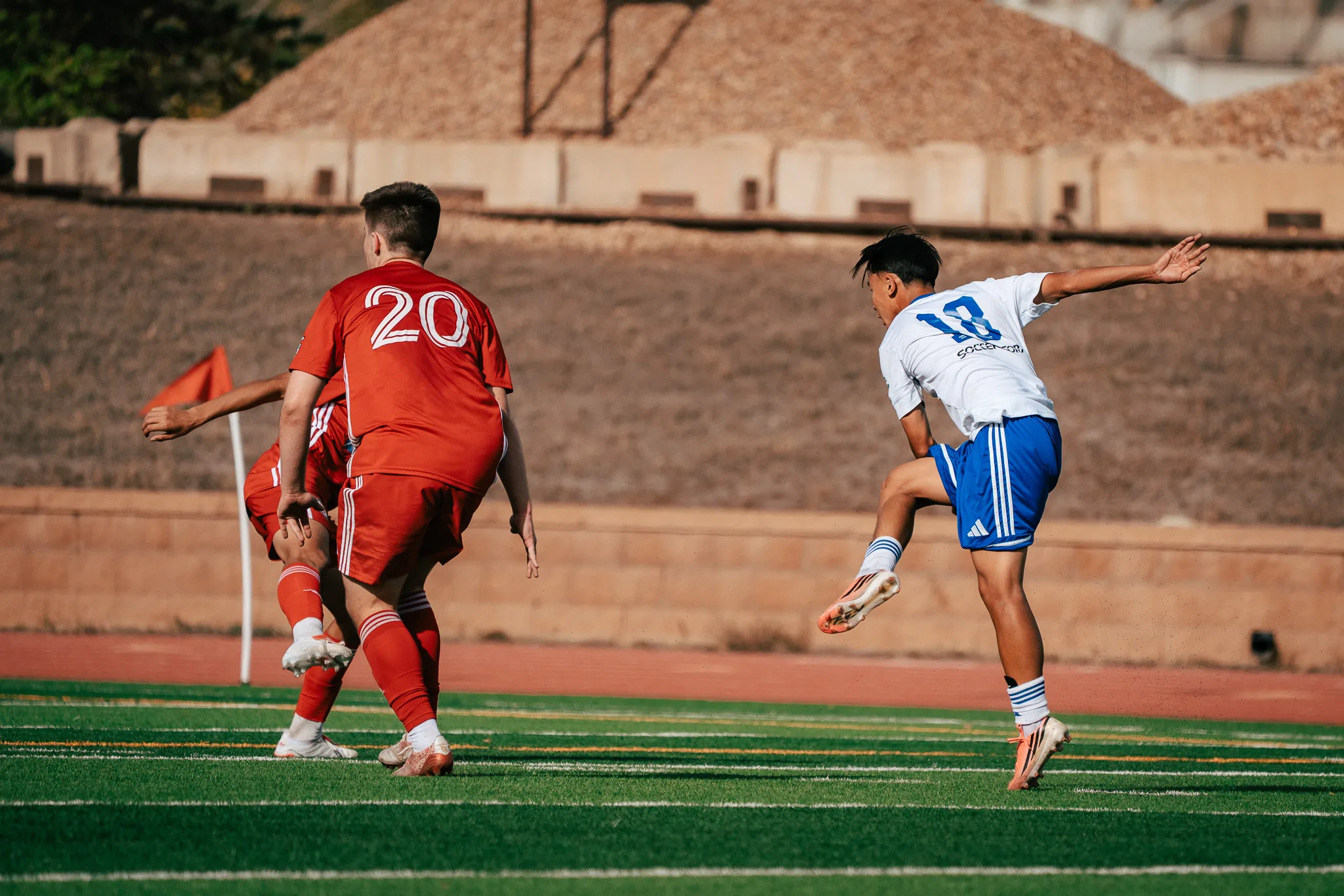 Albion SC Colorado number 18 swings through a kick in the air near the corner flag as AC River number 20 and a teammate turn away