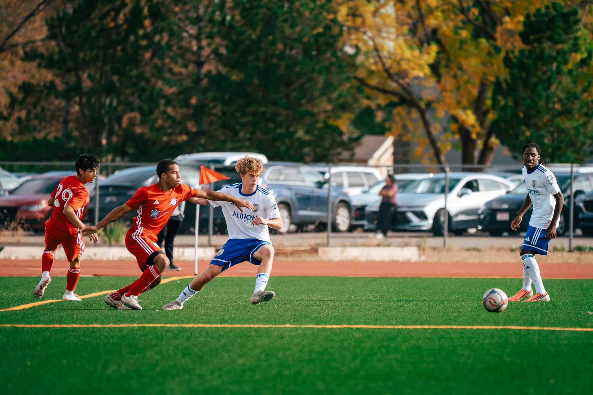 An Albion SC Colorado player with curly blond hair holds off two AC River players, number 29 one of them, as the ball rolls toward a teammate with long locs
