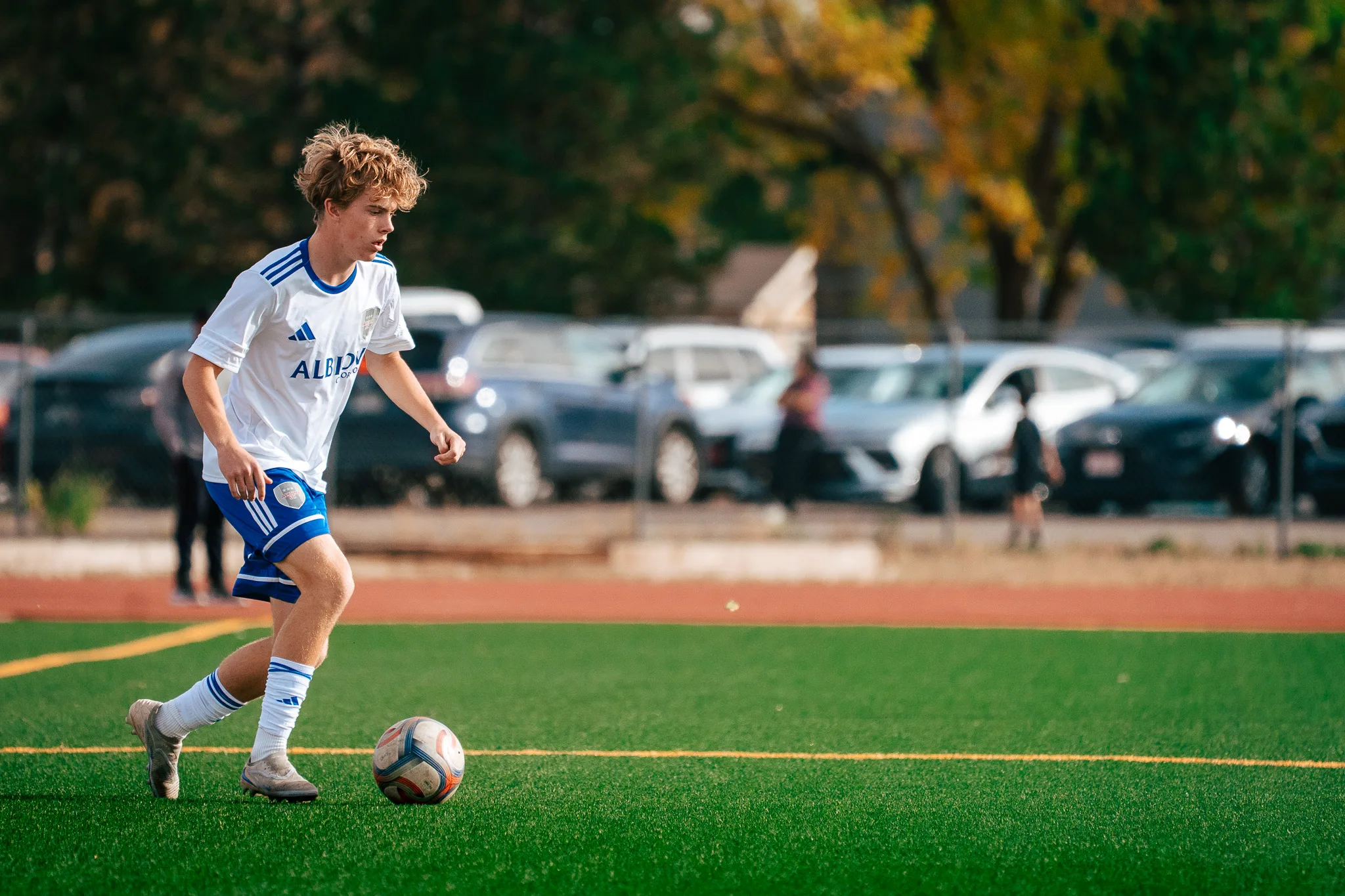 An Albion SC Colorado player with curly blond hair dribbles up the field, parked cars behind the fence