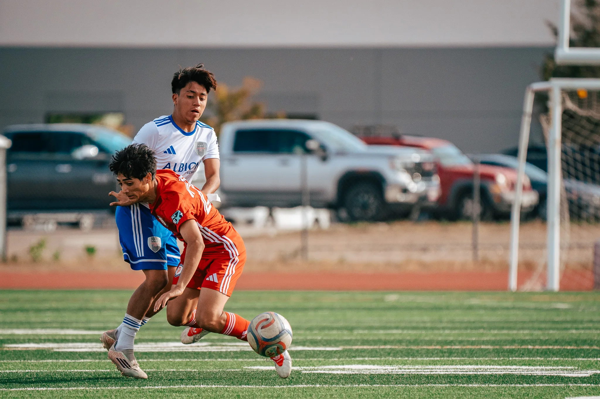 An AC River player leans over the ball as an Albion SC Colorado player presses from behind, parked trucks beyond the field
