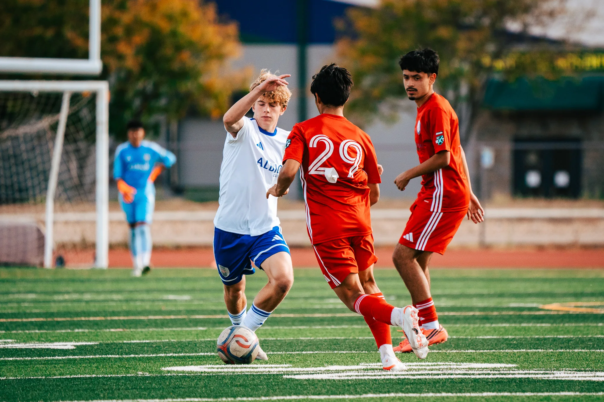 An Albion SC Colorado player with curly blond hair plays the ball with a hand at his head as AC River number 29 closes in, the Albion goalkeeper in light blue behind