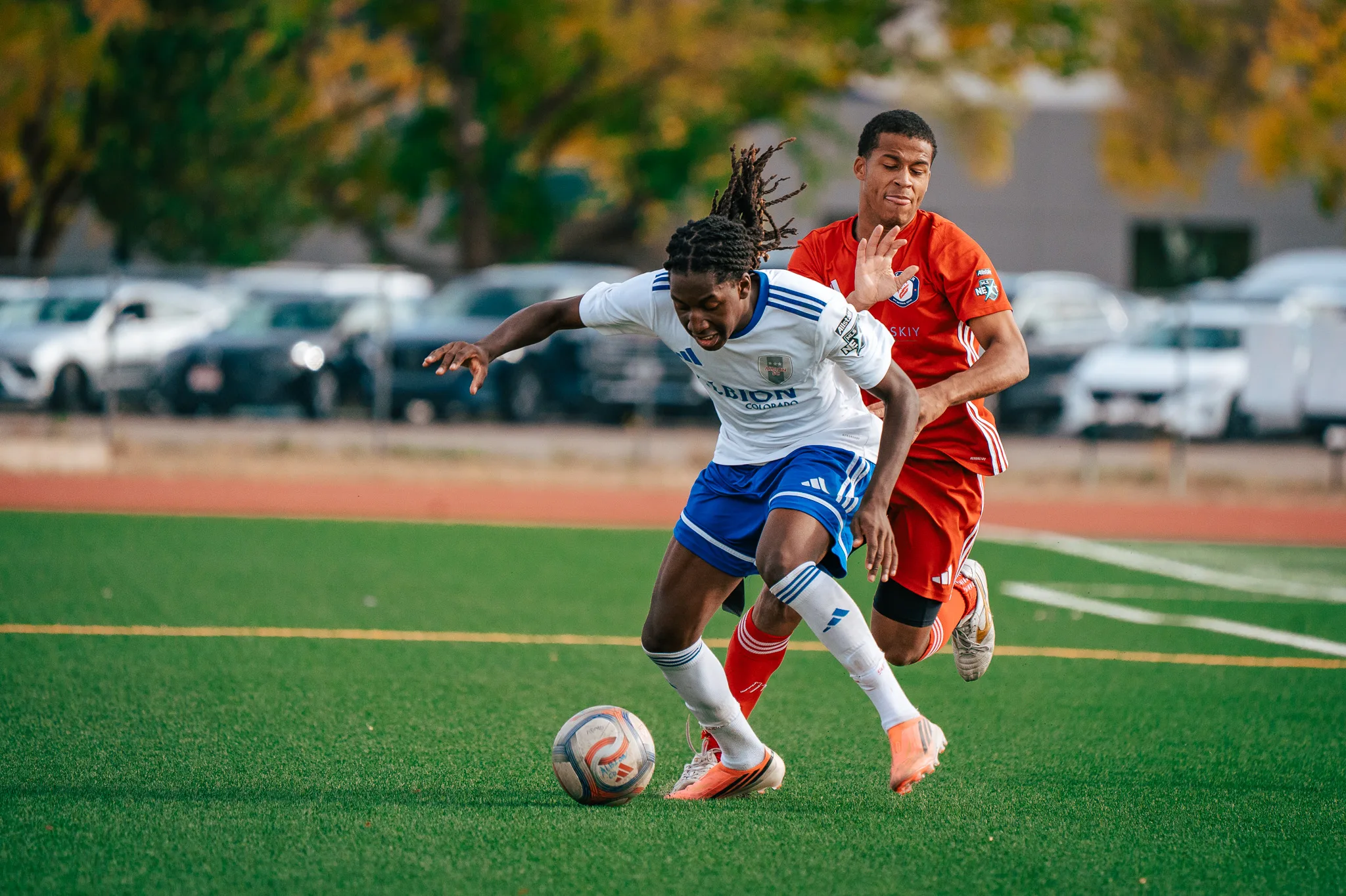 An Albion SC Colorado player with long locs drives past an AC River player reaching around him