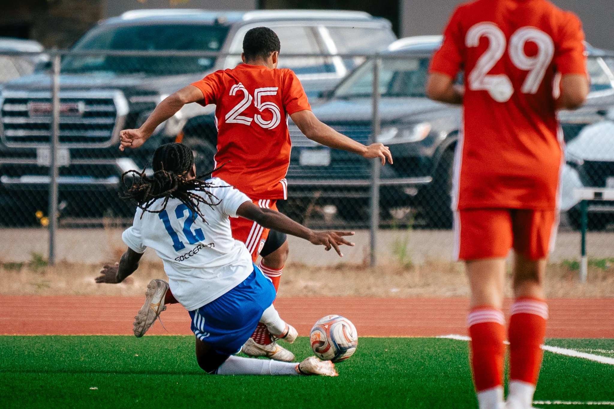 Albion SC Colorado number 12, his locs flying, slides in on the ball against AC River number 25 as AC River number 29 looks on