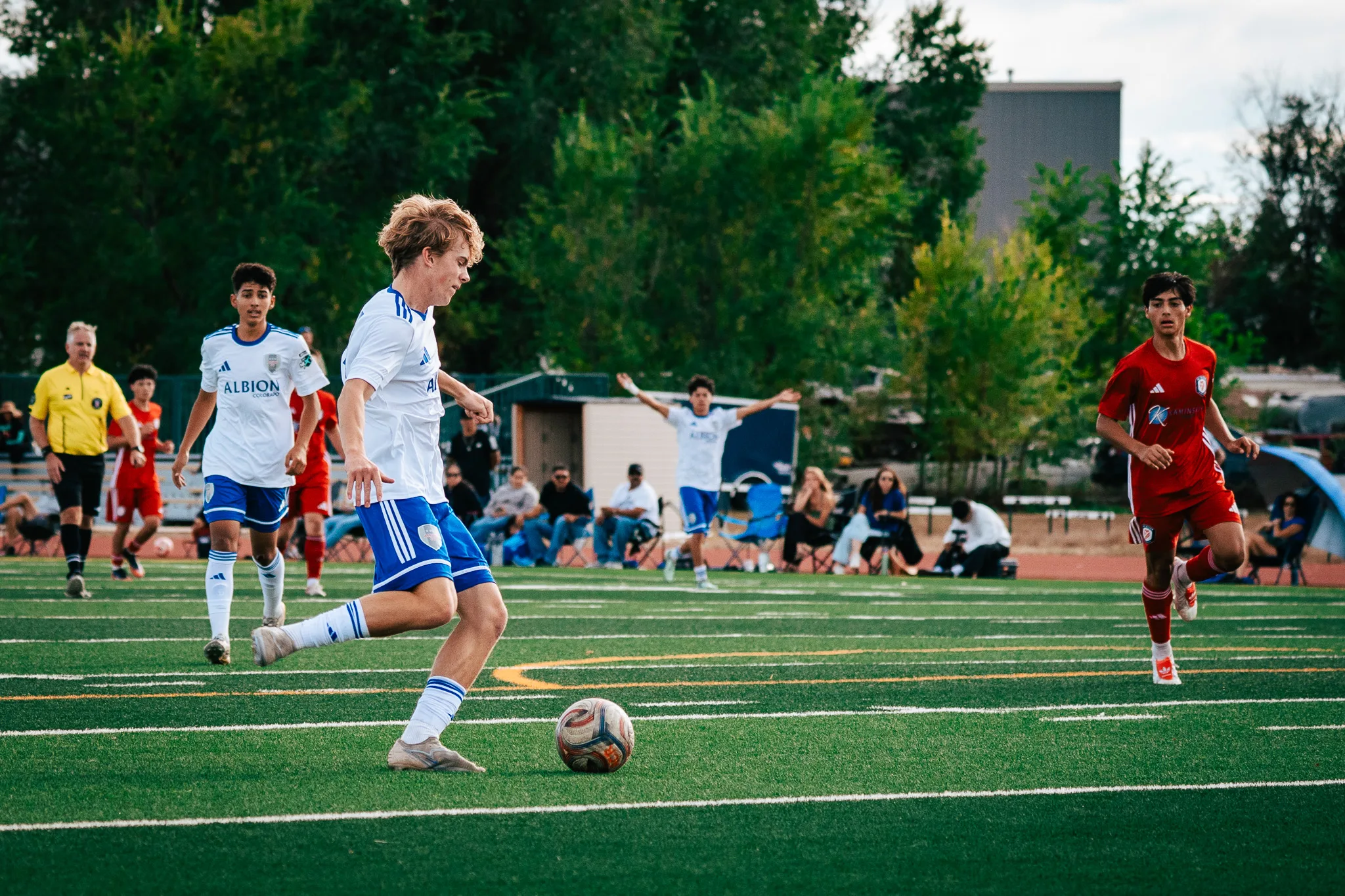 An Albion SC Colorado player with blond hair dribbles as teammates spread out behind him, one with both arms raised, and an AC River player closes from the right