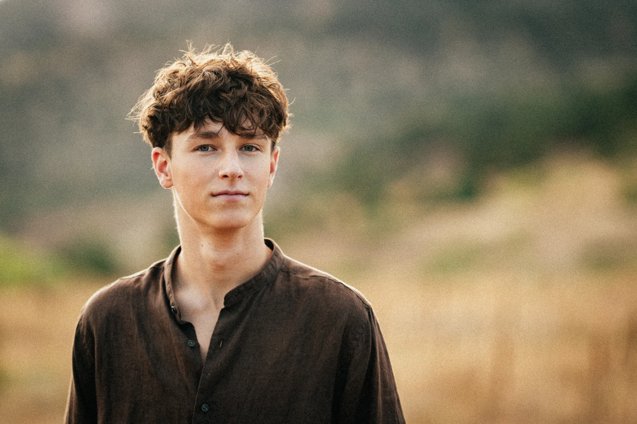 Arlo looks calmly into the camera in a brown linen shirt, a golden meadow blurred behind.