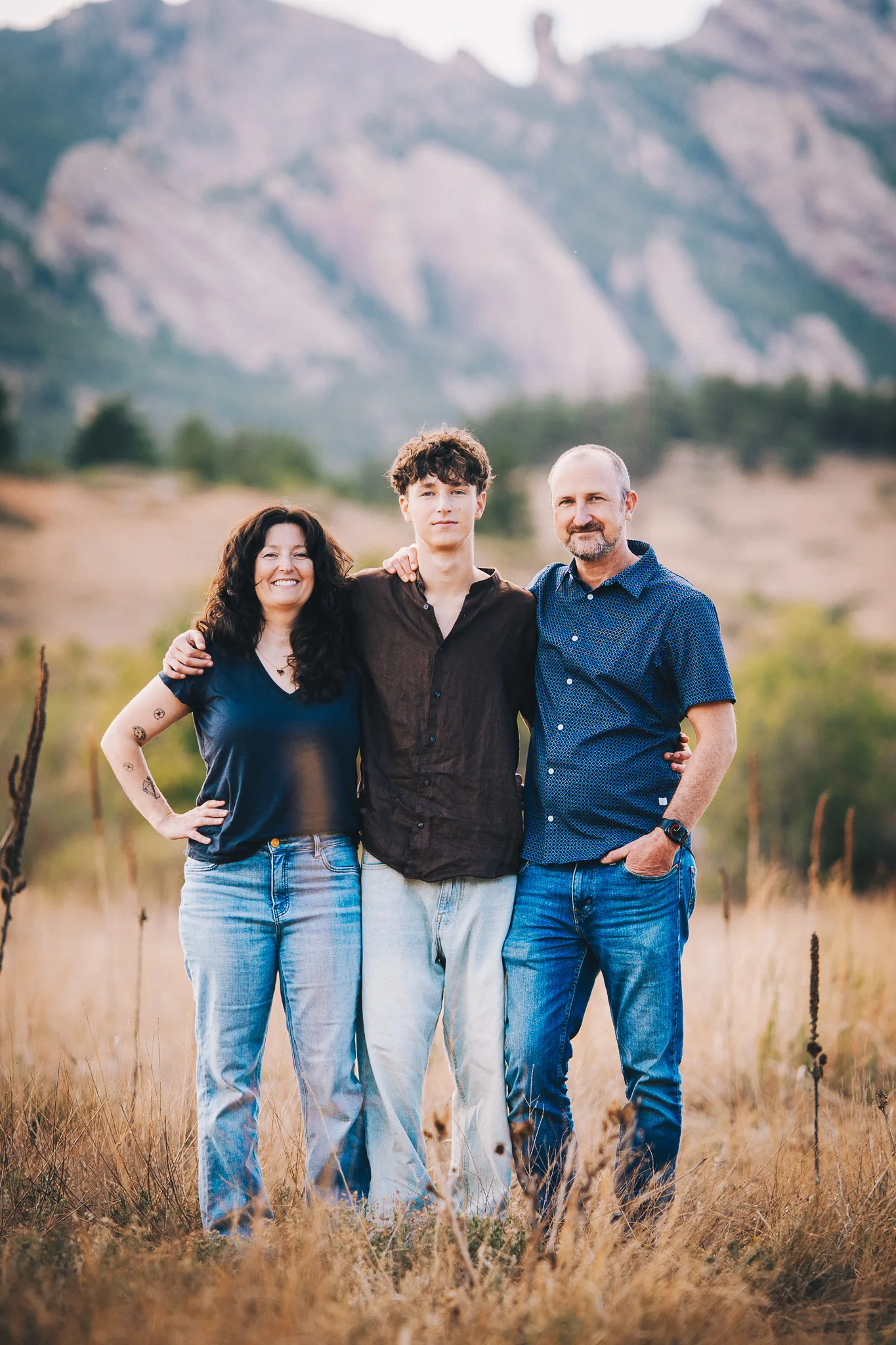Arlo stands between his mom, Amy, and his dad, Dusty, in tall grass with the Flatirons behind.