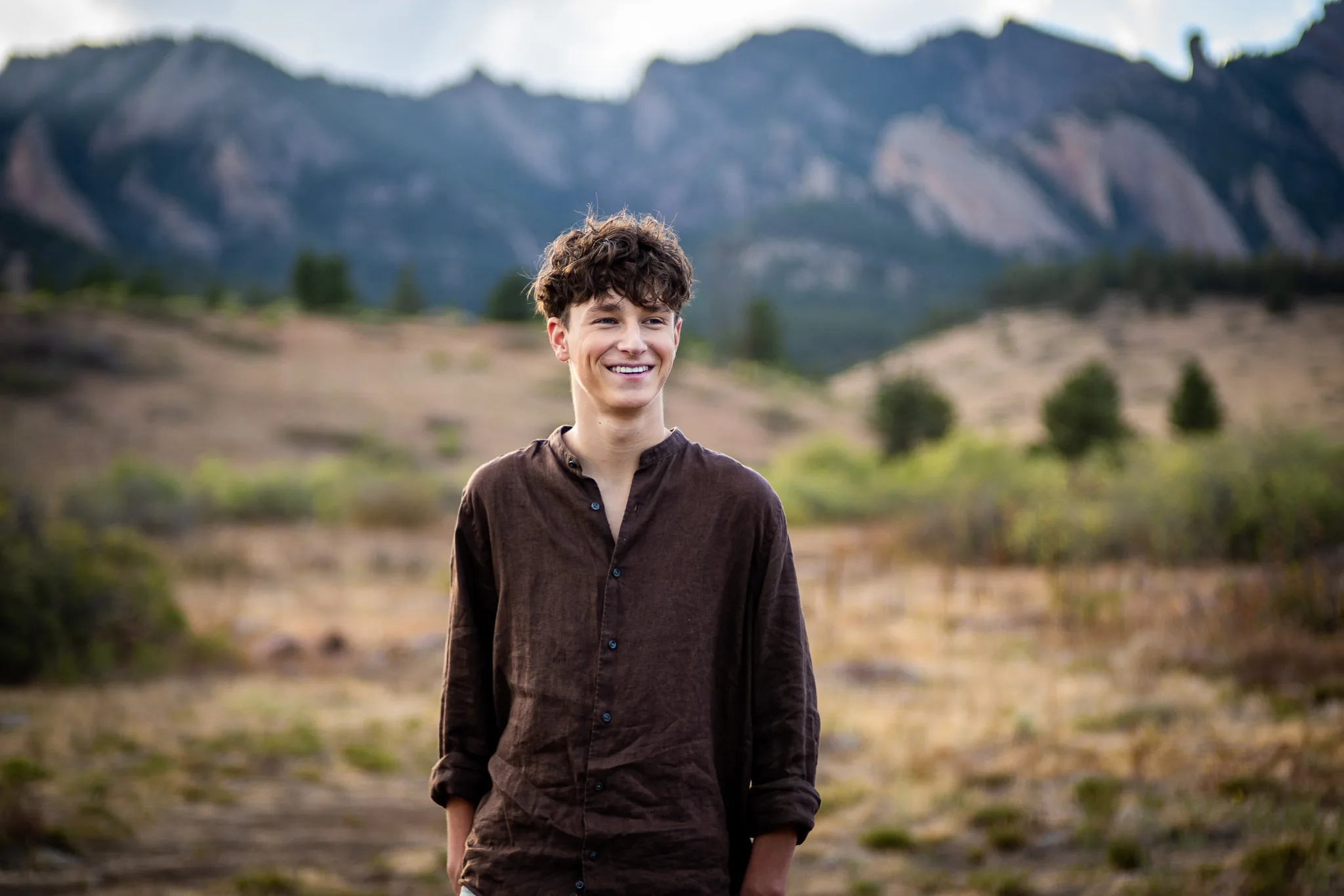 Arlo, in a brown linen shirt, smiles in an open meadow below the Flatirons at dusk.