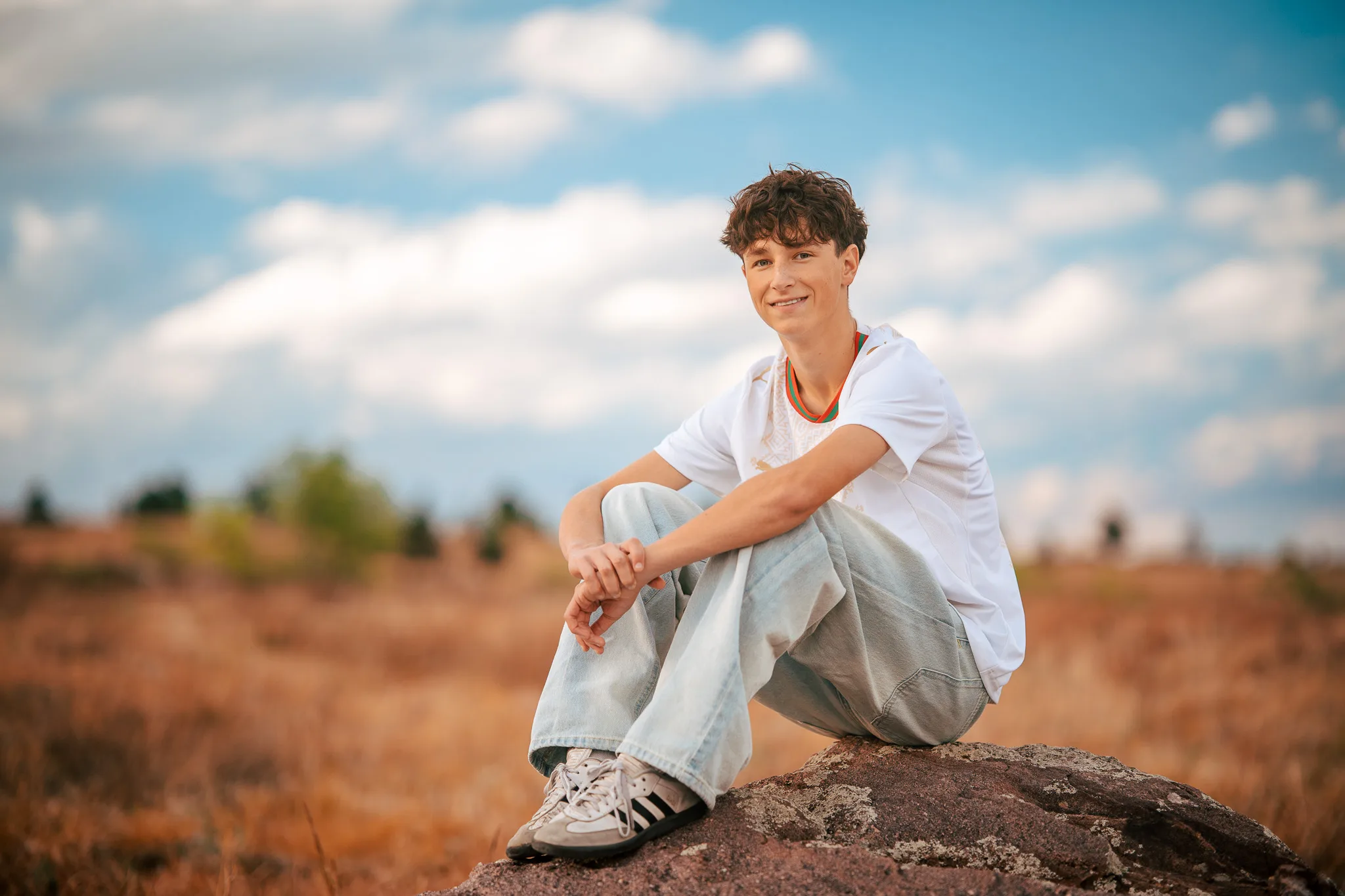 Arlo smiles from a boulder in a golden meadow under a blue sky and white clouds.
