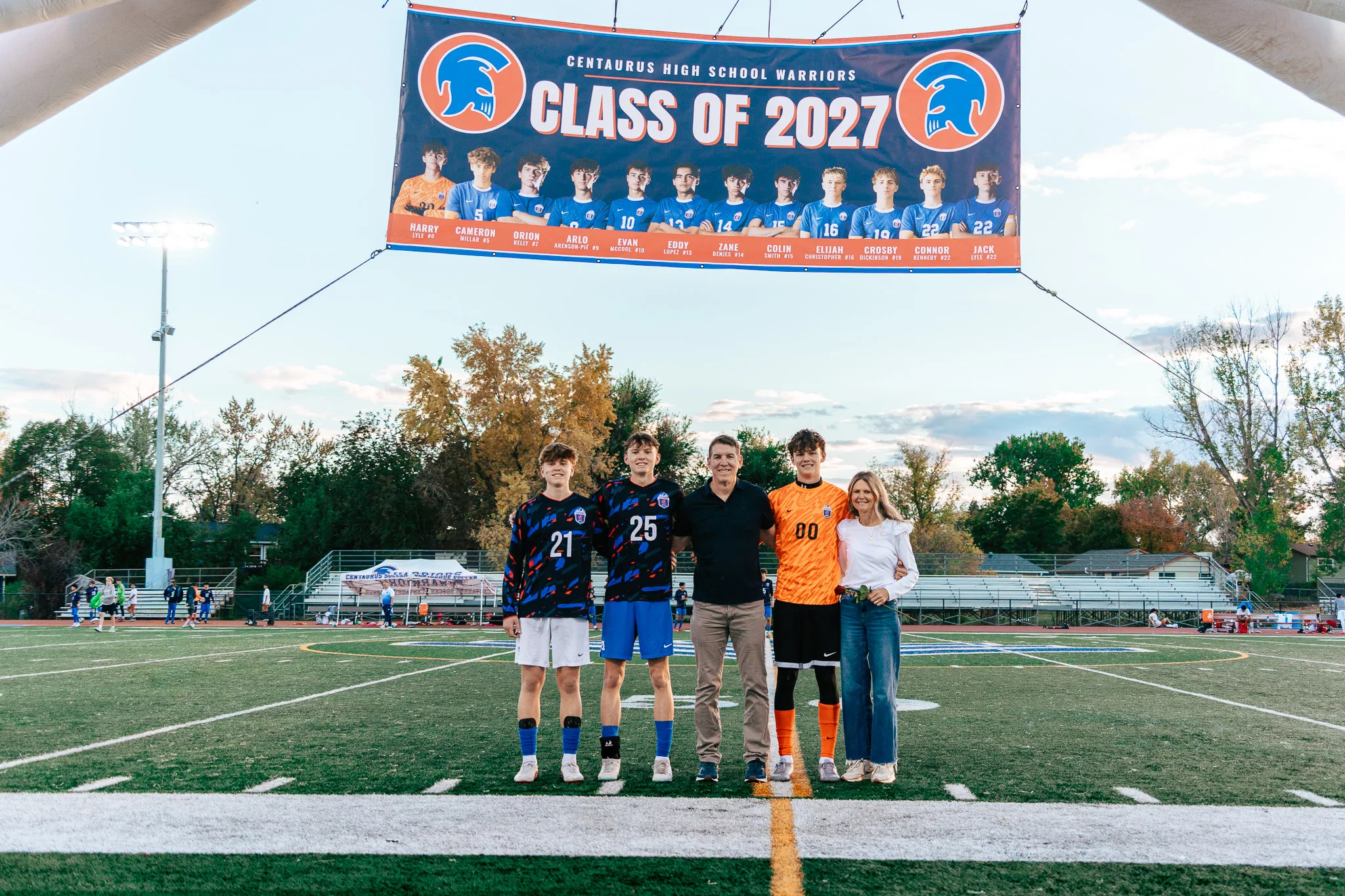 Under a Class of 2027 banner hung above the field, the Centaurus goalkeeper in orange stands with a man in a black polo, a woman in white and two teammates in numbers 21 and 25, autumn trees and bleachers behind