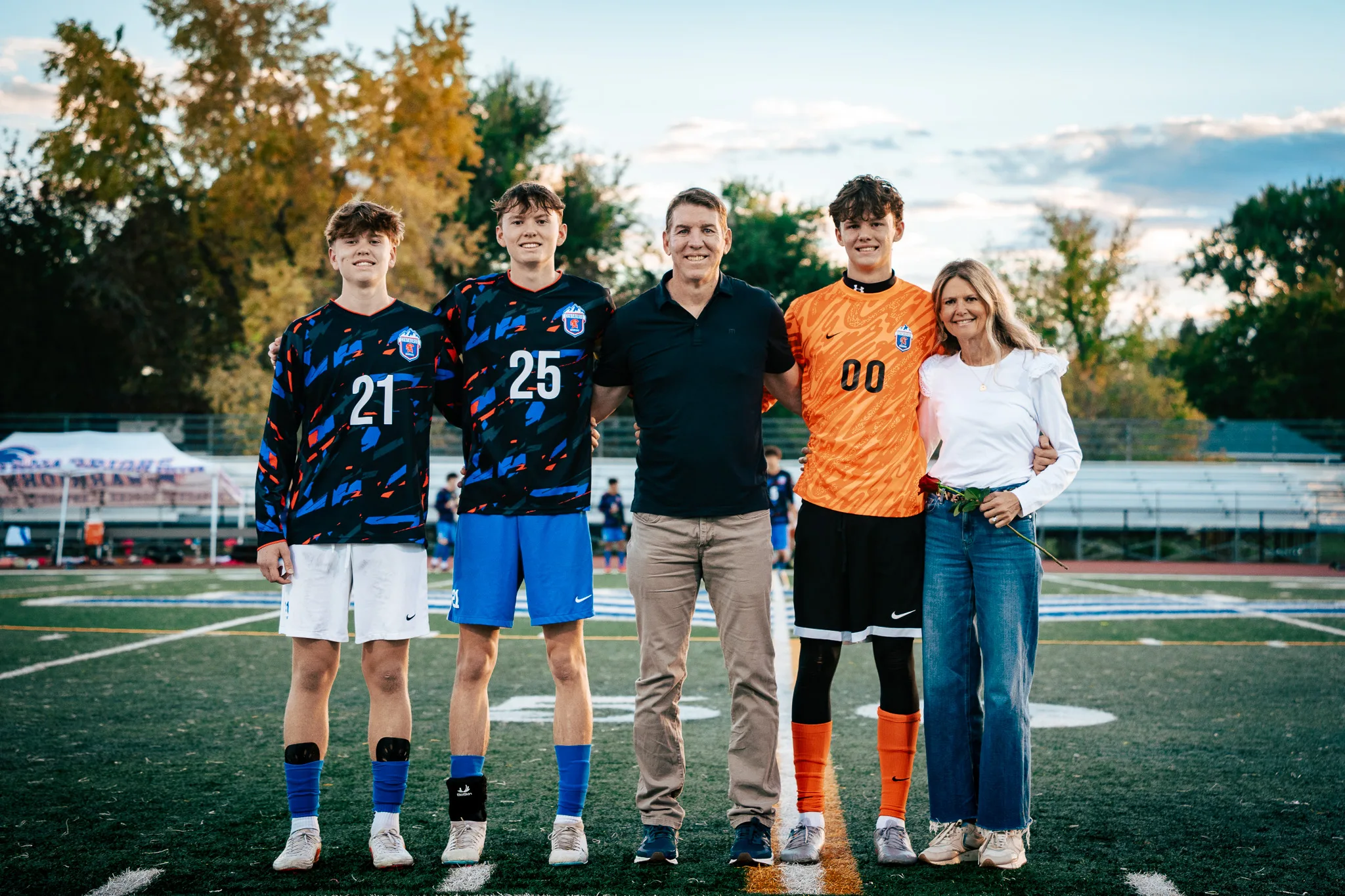 Centaurus numbers 21 and 25, a man in a black polo, the goalkeeper in orange and a woman in white holding a red rose pose arm in arm at midfield, shown full length with autumn trees behind