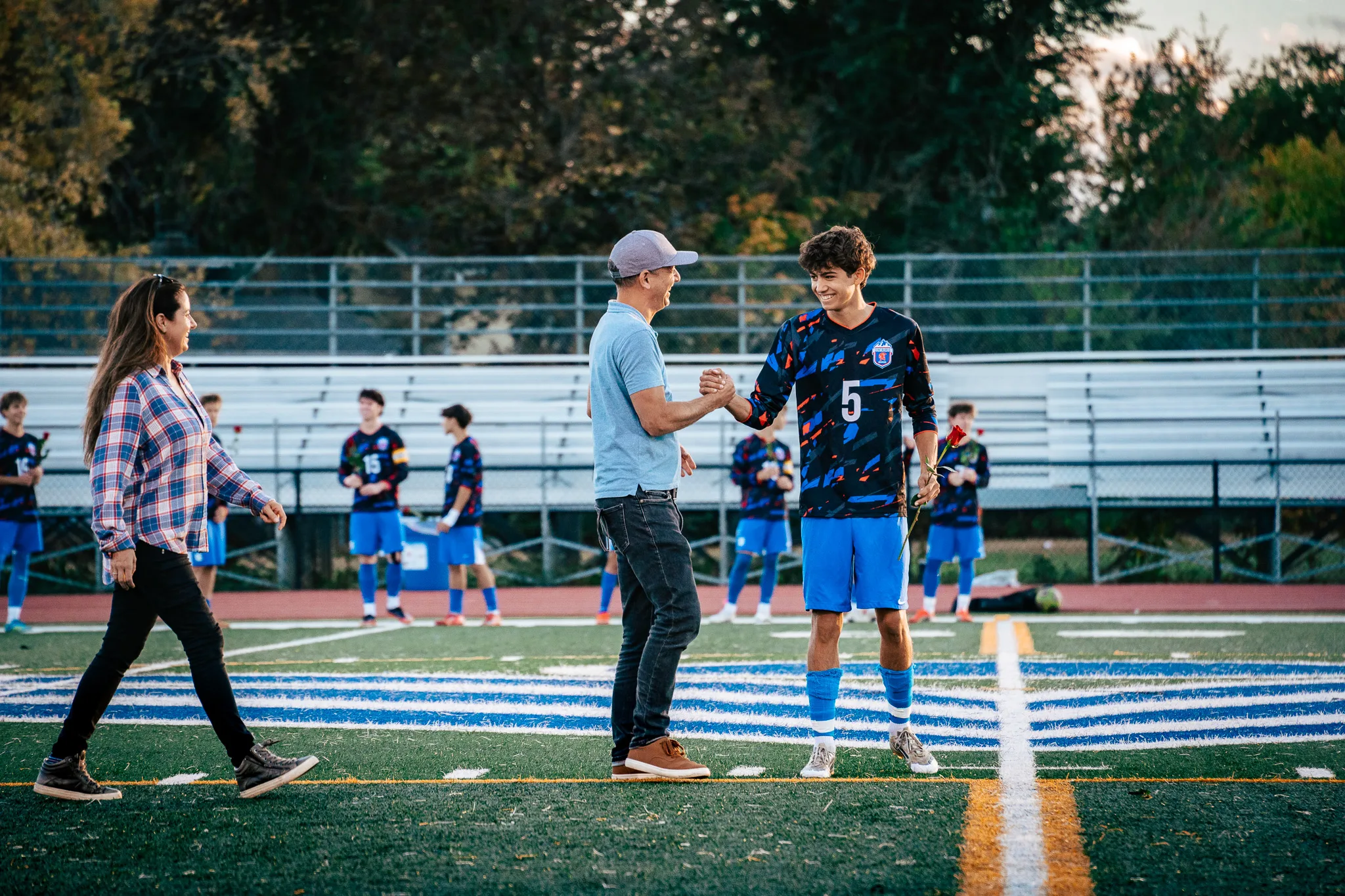 Centaurus number 5, a red rose in his hand, shakes hands with a man in a gray cap and light blue polo as a woman in a plaid shirt walks past, teammates waiting along the sideline behind