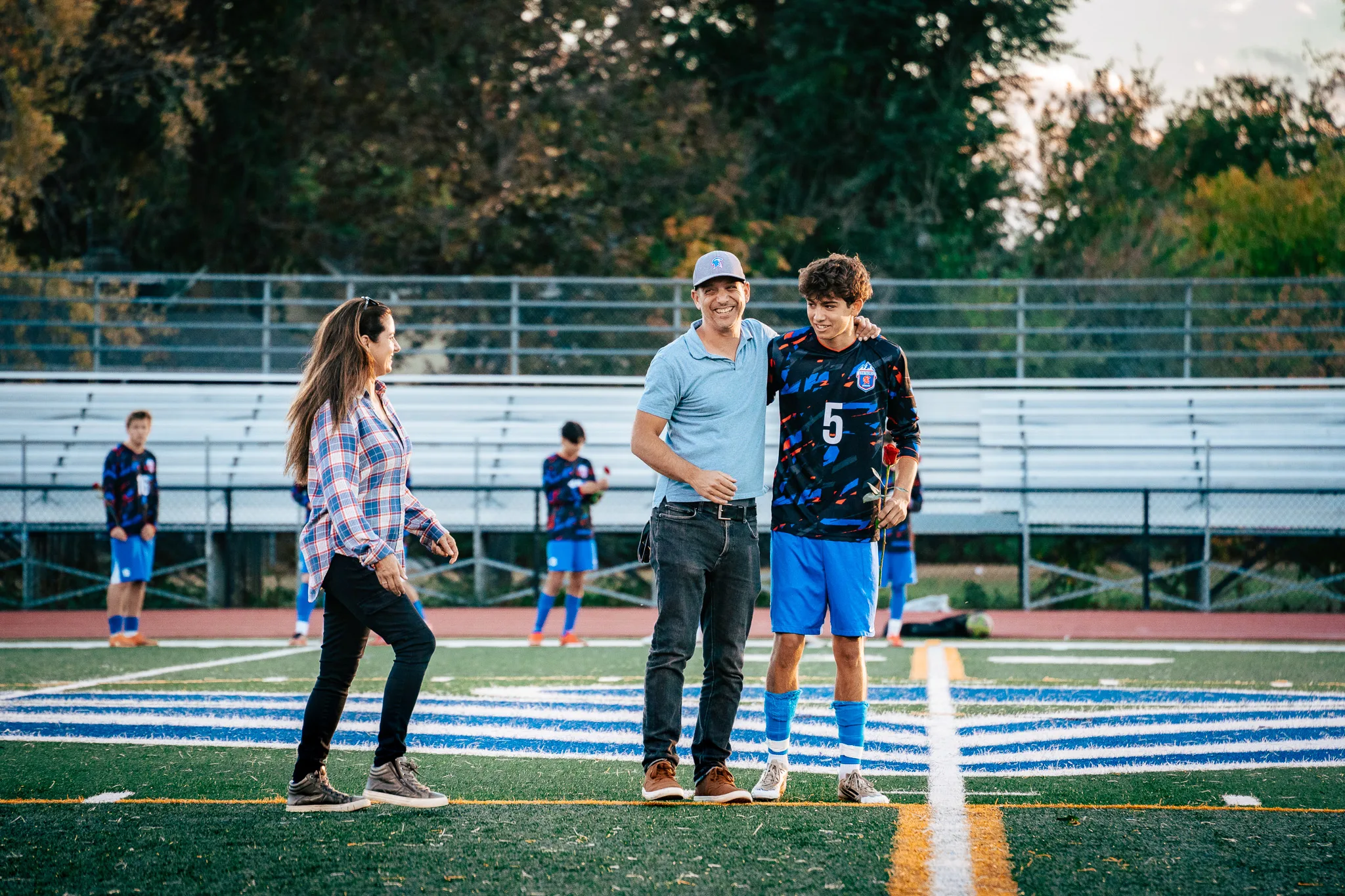 Centaurus number 5 holds a red rose as a laughing man in a gray cap puts an arm around his shoulder and a woman in a plaid shirt looks on, teammates standing along the bleachers behind