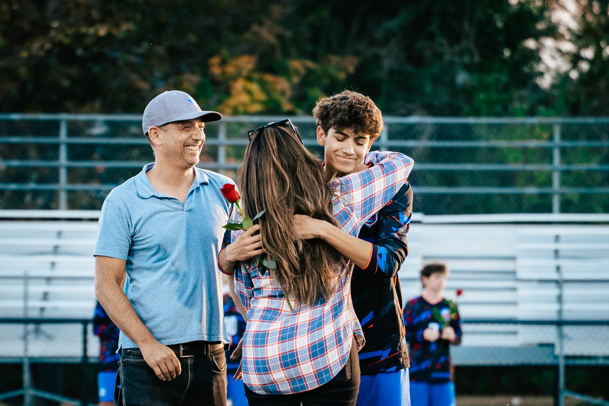 A woman in a plaid shirt hugs a smiling Centaurus player, his curly hair showing over her shoulder, as a man in a gray cap laughs beside them, a red rose between them
