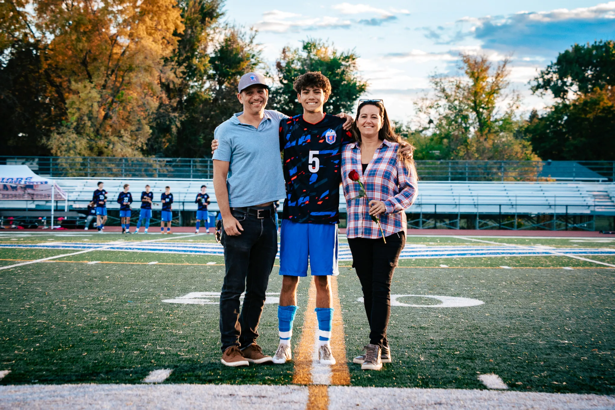 Centaurus number 5 smiles at midfield with his arms around a man in a gray cap and a woman in a plaid shirt holding a red rose, autumn trees and teammates along the sideline behind them