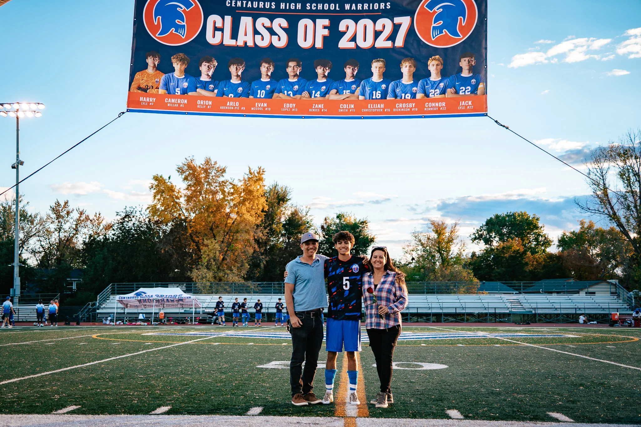 Centaurus number 5 poses at midfield with a man in a gray cap and a woman in a plaid shirt, small beneath a Class of 2027 banner, with autumn trees and the bleachers behind