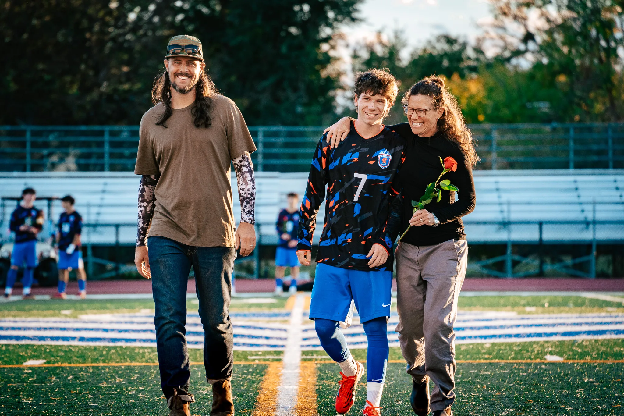 Centaurus number 7 smiles as he walks onto the field between a bearded man with long hair in a cap and a woman in glasses holding a red rose, her arm around him, teammates waiting along the sideline behind