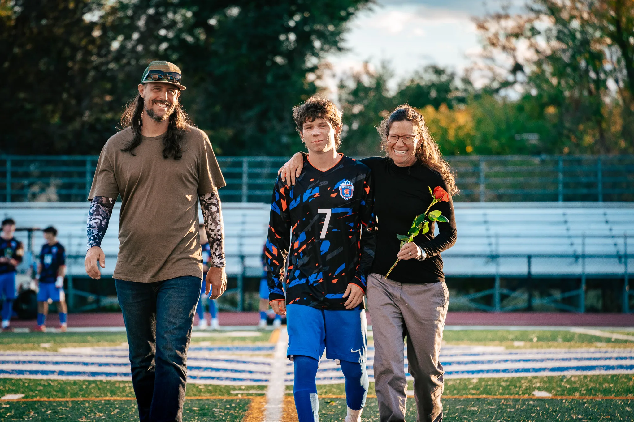 Centaurus number 7 walks arm in arm between a smiling bearded man with long hair in a cap and a woman in glasses holding a red rose, the midfield stripes under their feet
