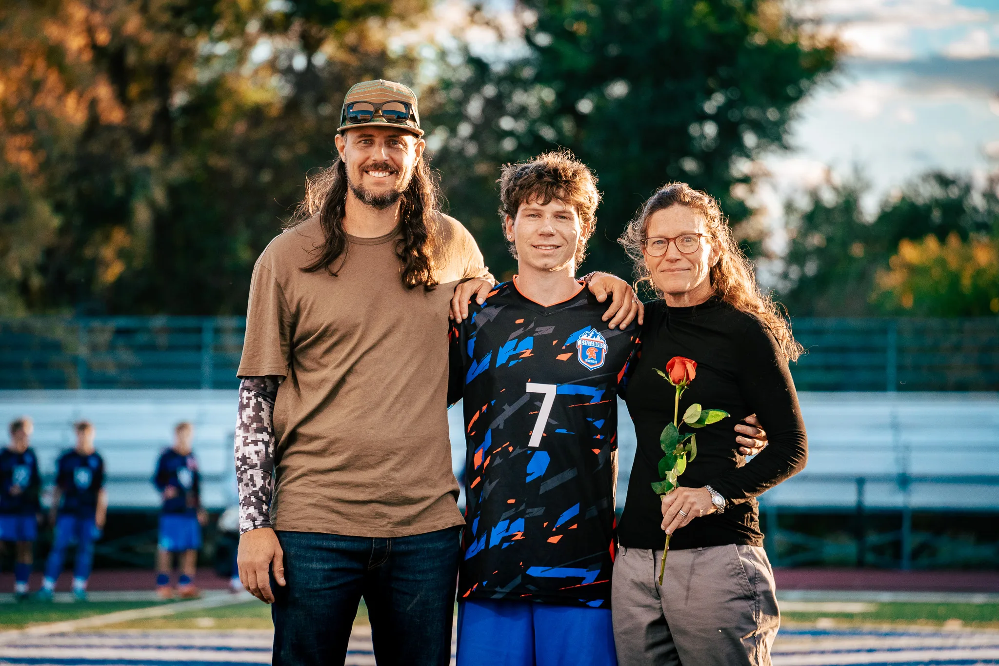 Centaurus number 7 poses with his arms around a smiling bearded man in a cap and a woman in glasses holding a red rose, autumn trees blurred behind