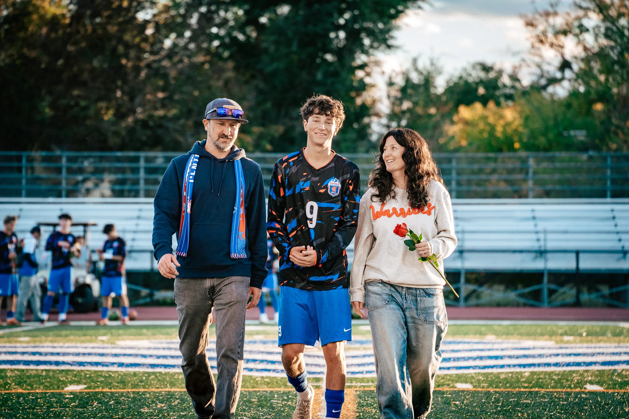 Centaurus number 9 walks onto the field between a bearded man in a navy hoodie and a blue and orange scarf and a woman in a Warriors sweatshirt holding a red rose, all three smiling