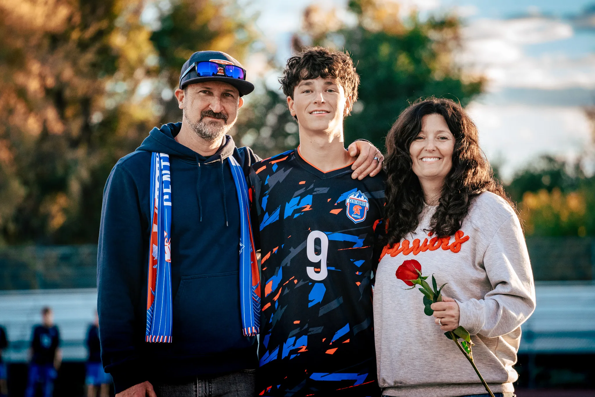 Close up, Centaurus number 9 smiles between a bearded man in a cap and a blue and orange scarf and a woman in a Warriors sweatshirt holding a red rose