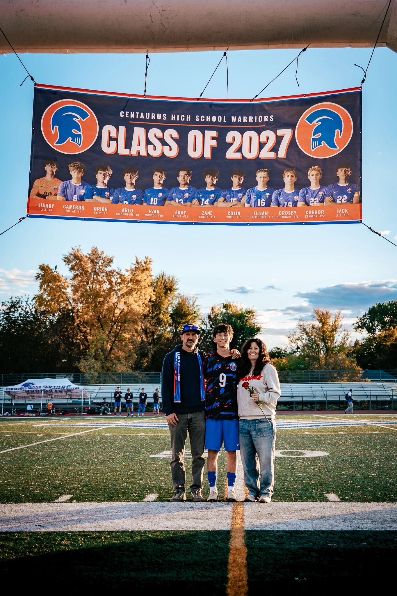 Beneath a Class of 2027 banner strung over the field, Centaurus number 9 stands with a man in a blue and orange scarf and a woman in a Warriors sweatshirt holding a red rose, autumn trees and the bleachers behind them