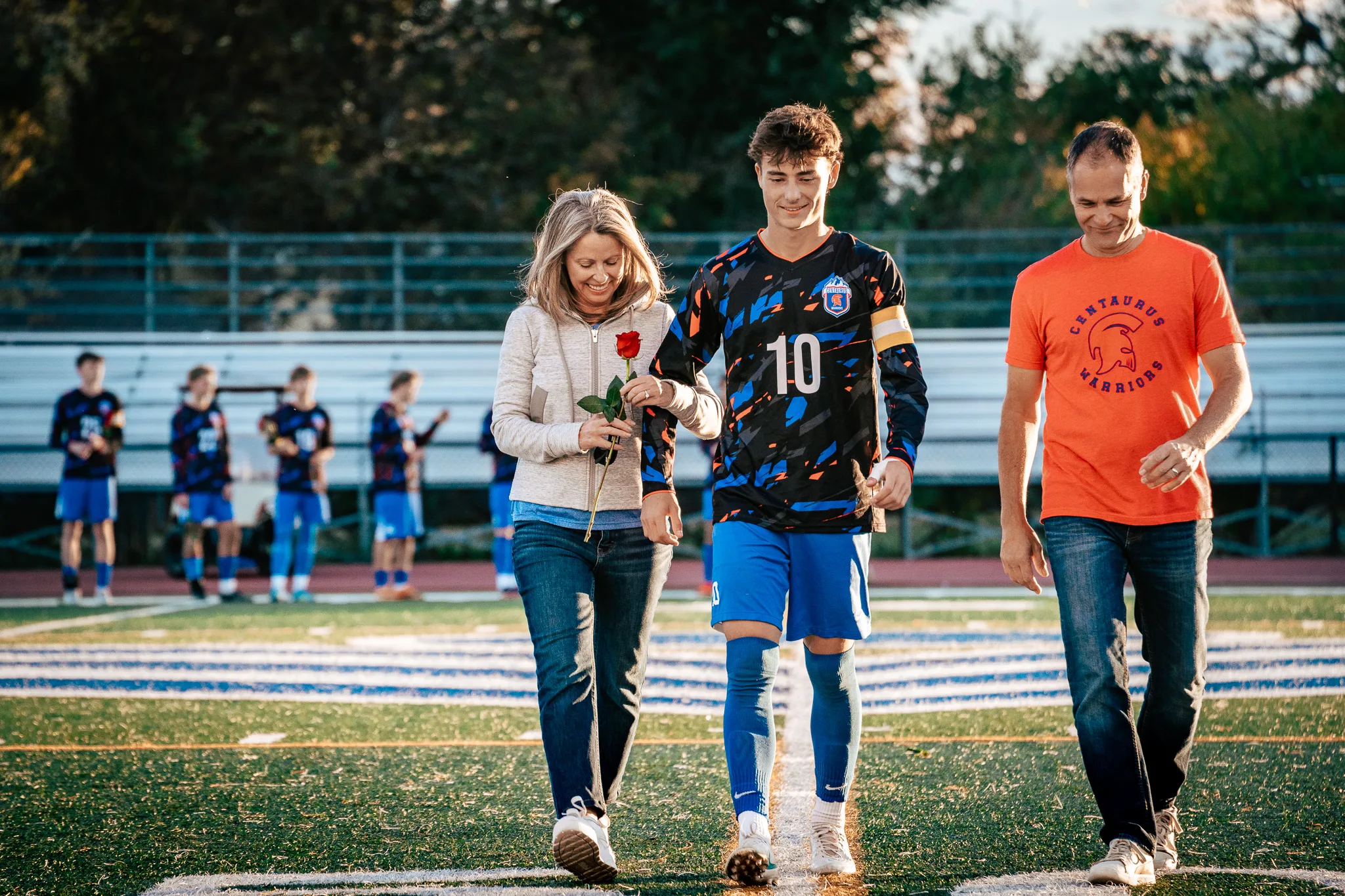 Centaurus number 10, in a captain's armband, walks onto the field between a smiling woman in a cream jacket holding a red rose and a man in an orange Centaurus Warriors shirt