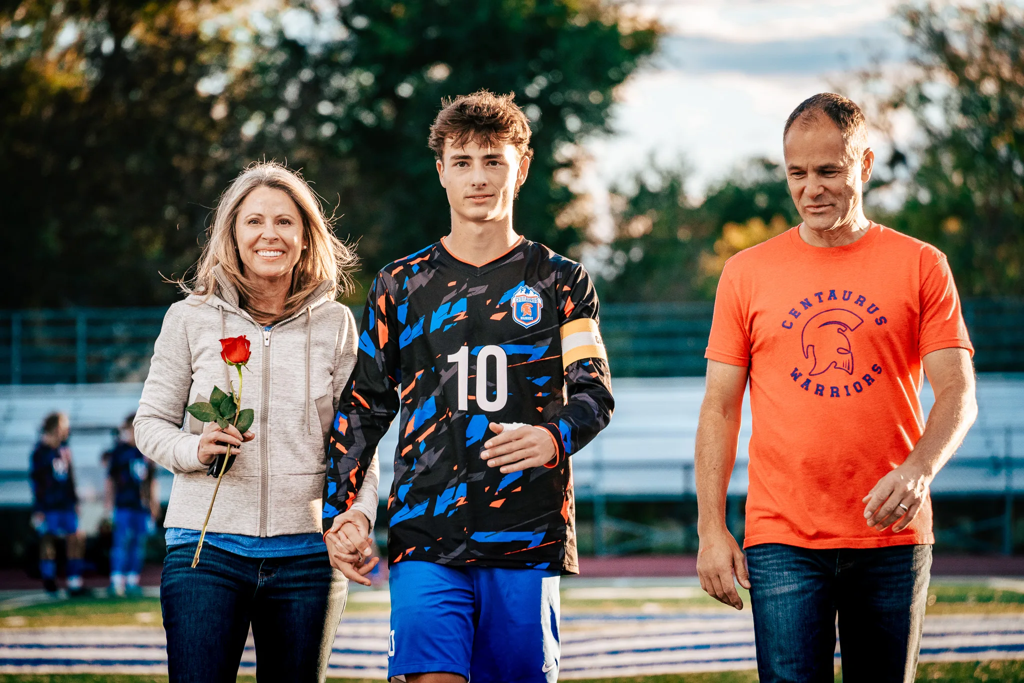 Close up, Centaurus number 10 in a captain's armband holds hands with a smiling woman holding a red rose as a man in an orange Centaurus Warriors shirt walks beside them