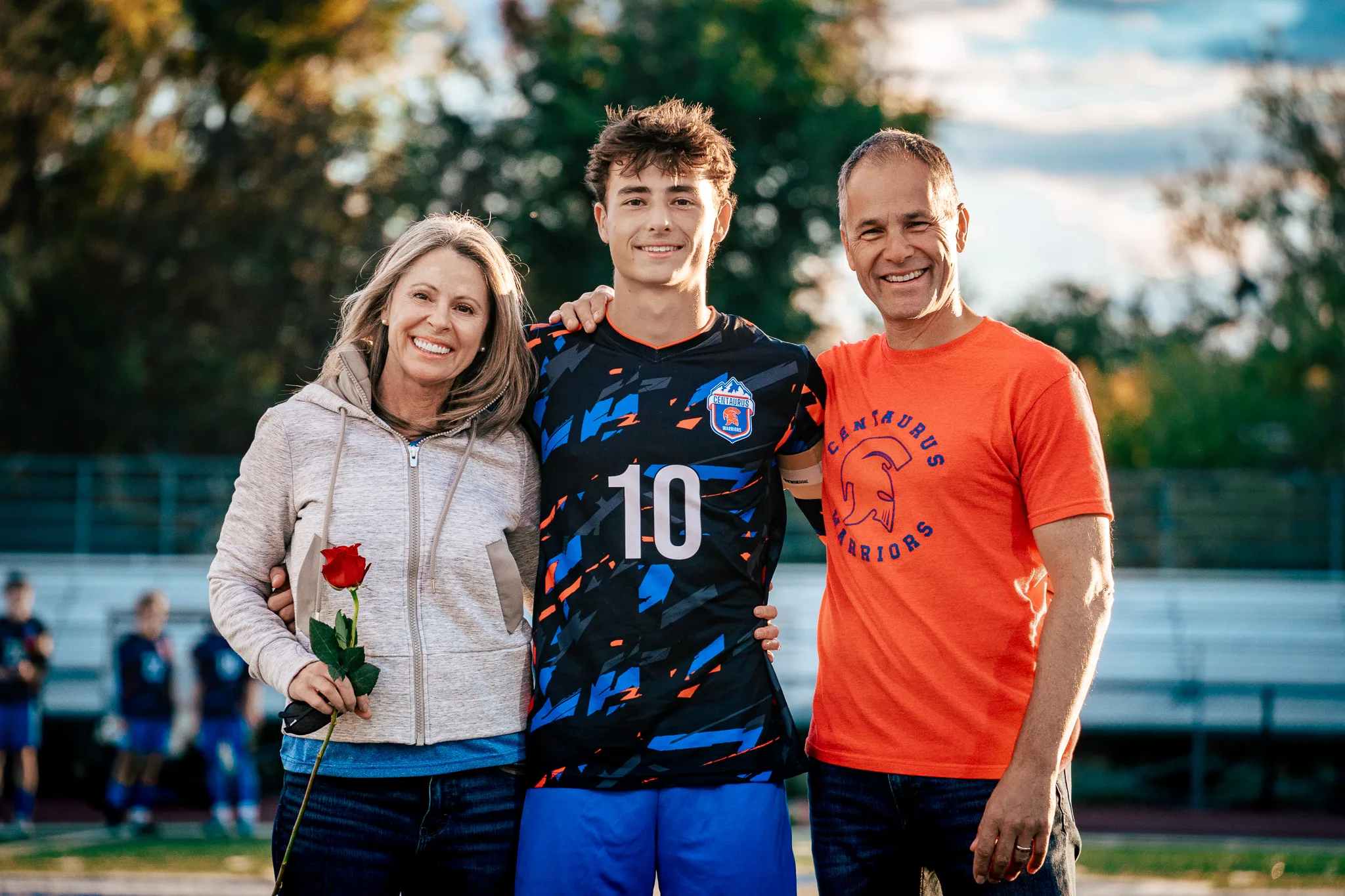 Centaurus number 10 poses with his arms around a smiling woman in a cream jacket holding a red rose and a man in an orange Centaurus Warriors shirt, autumn trees blurred behind