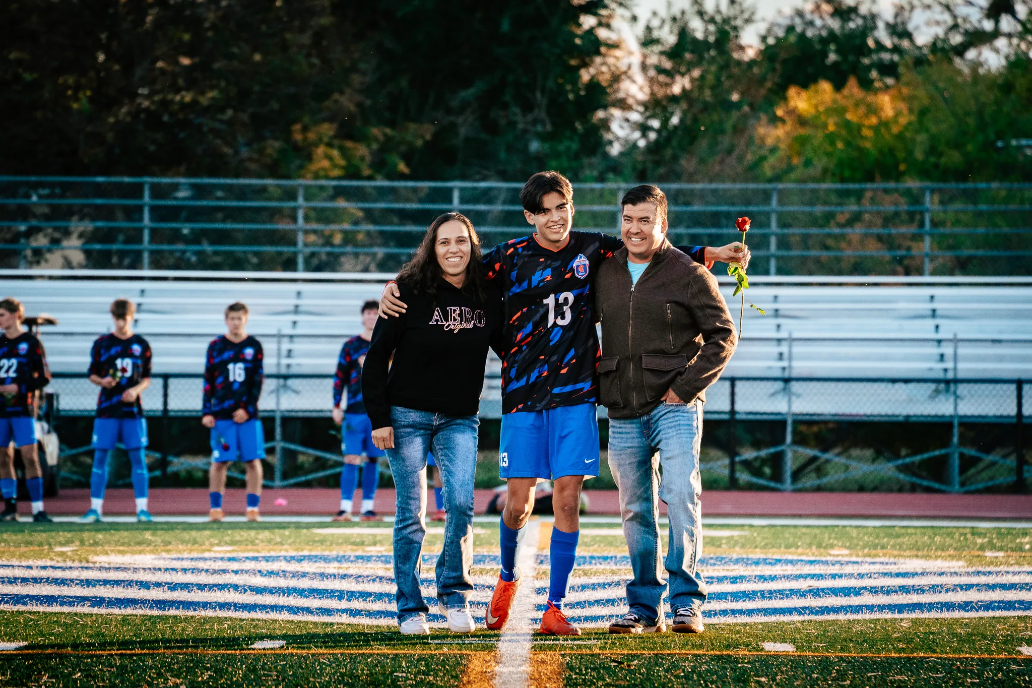 Centaurus number 13 walks onto the field arm in arm with a woman in a black sweatshirt and a man in a brown jacket who holds a red rose up behind him, teammates lined up along the sideline