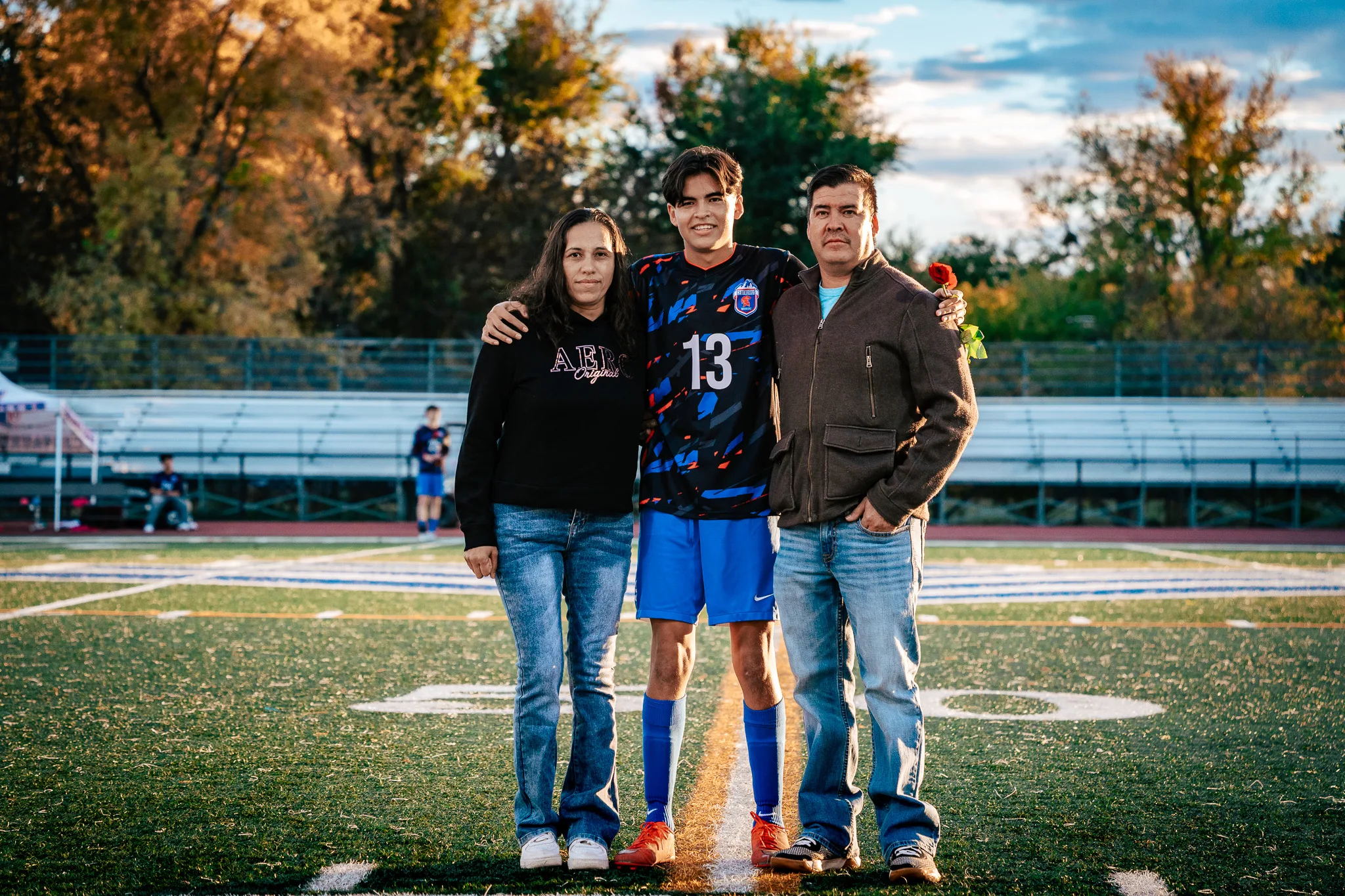 Centaurus number 13 poses at midfield with a woman in a black sweatshirt and a man in a brown jacket, a red rose in the man's hand, autumn trees and the bleachers behind